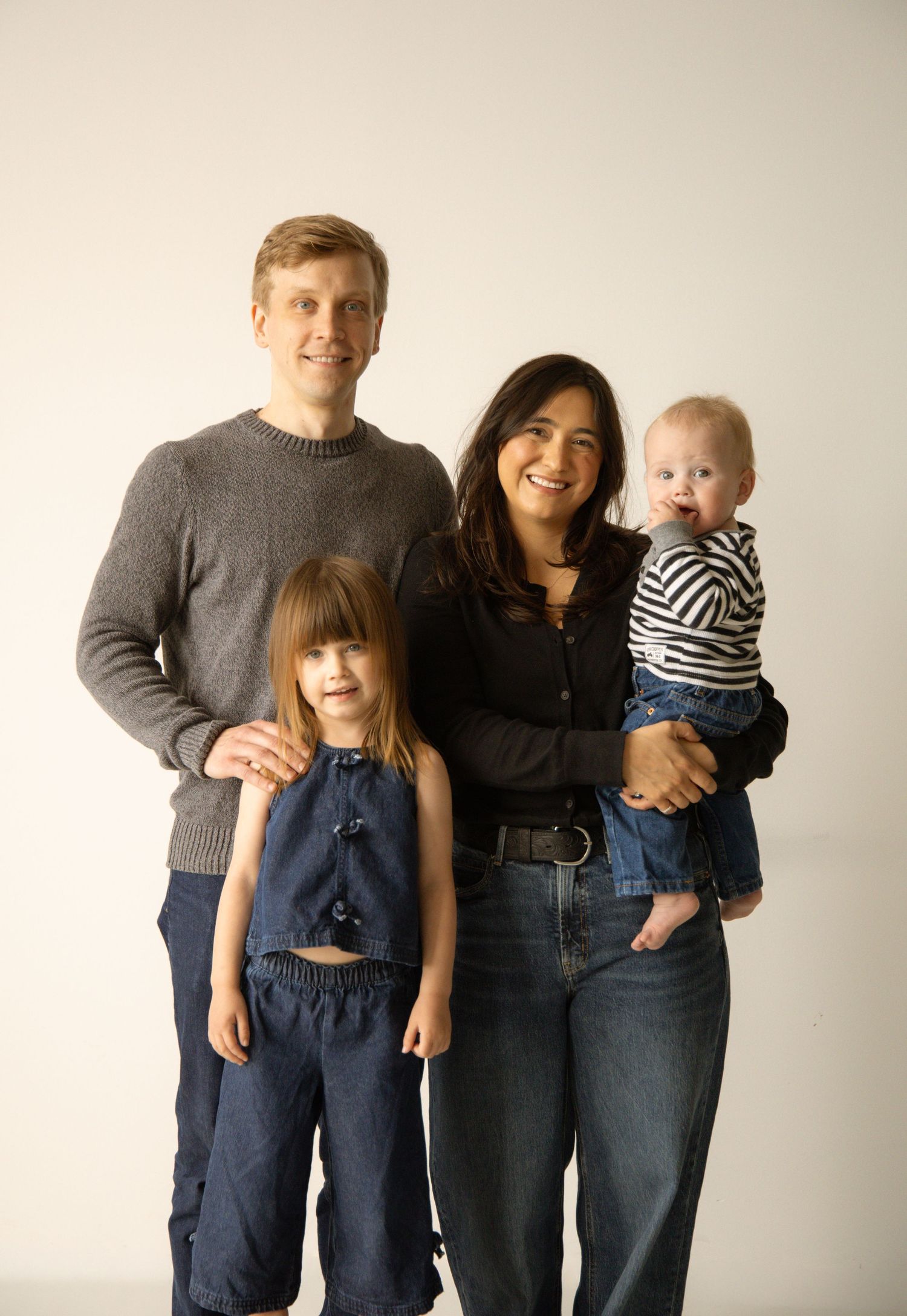 A smiling family of four poses together, with parents holding a baby in stripes and a young girl in denim overalls.