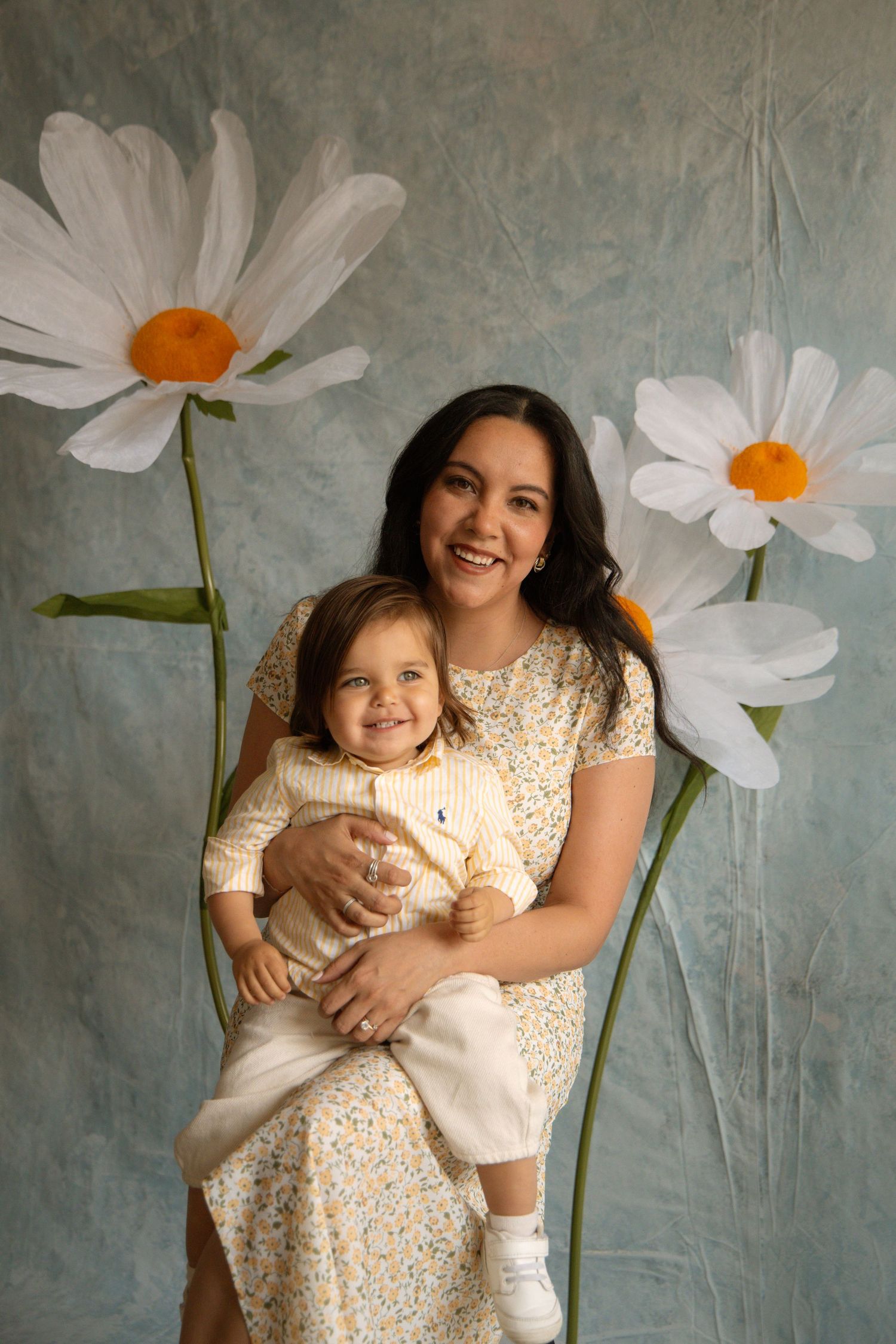 A smiling woman holds a happy toddler on her lap in front of a blue backdrop with large painted daisy flowers.