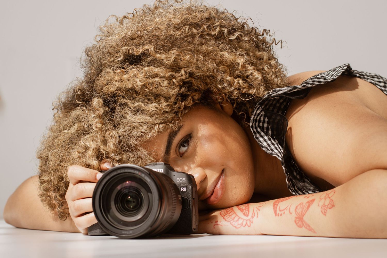 Woman with curly hair resting her head on a professional camera against a light background.