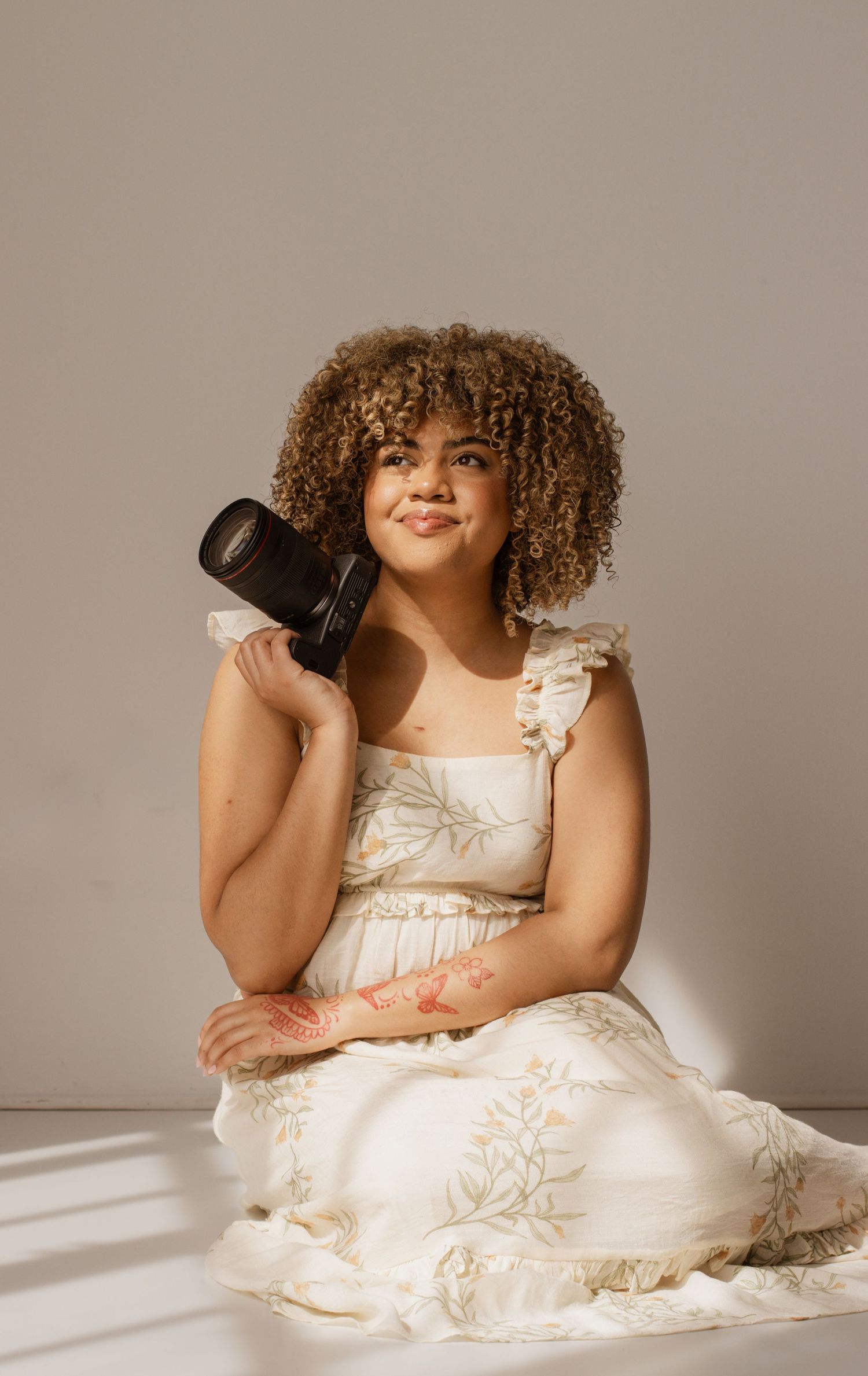 A young woman with curly hair in a white floral dress holds a DSLR camera while sitting and smiling softly.