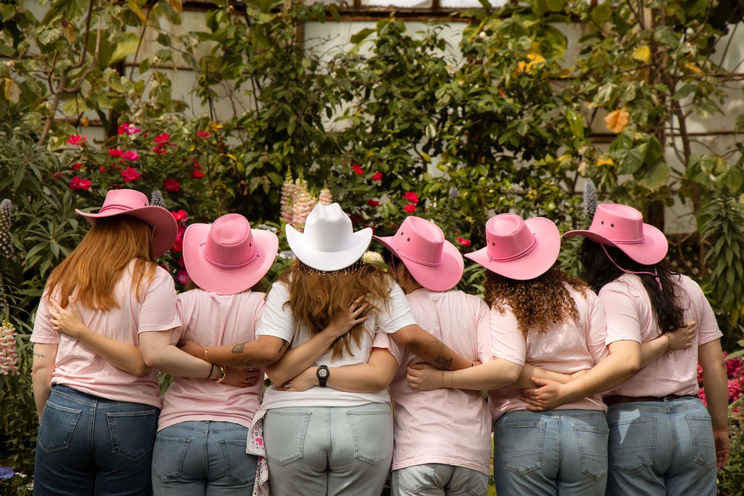 Six women in pink cowboy hats hugging in a row, viewed from behind, standing in front of lush garden greenery.