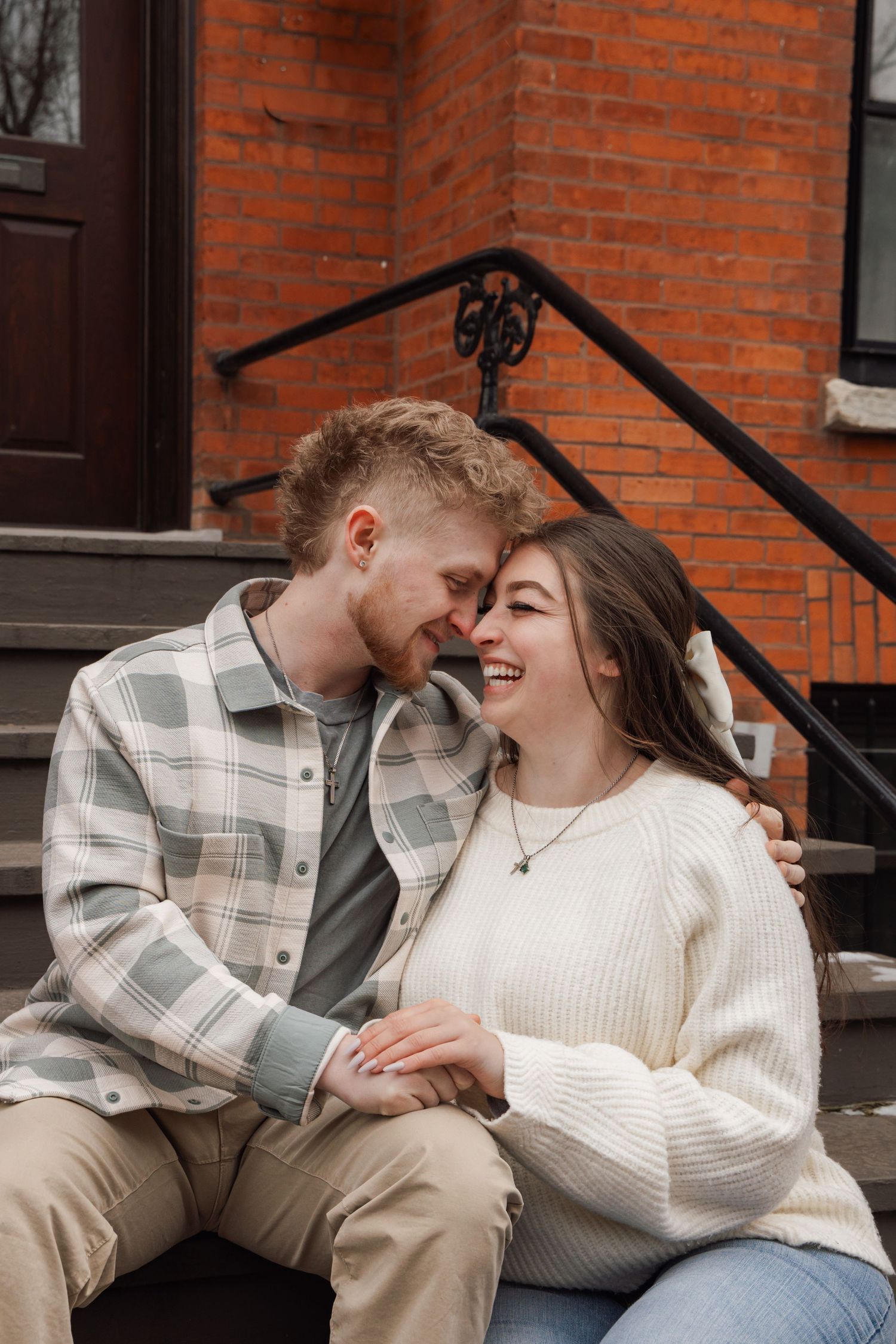 A smiling couple sitting on brick building steps, touching foreheads and holding hands in a cozy, romantic outdoor moment.