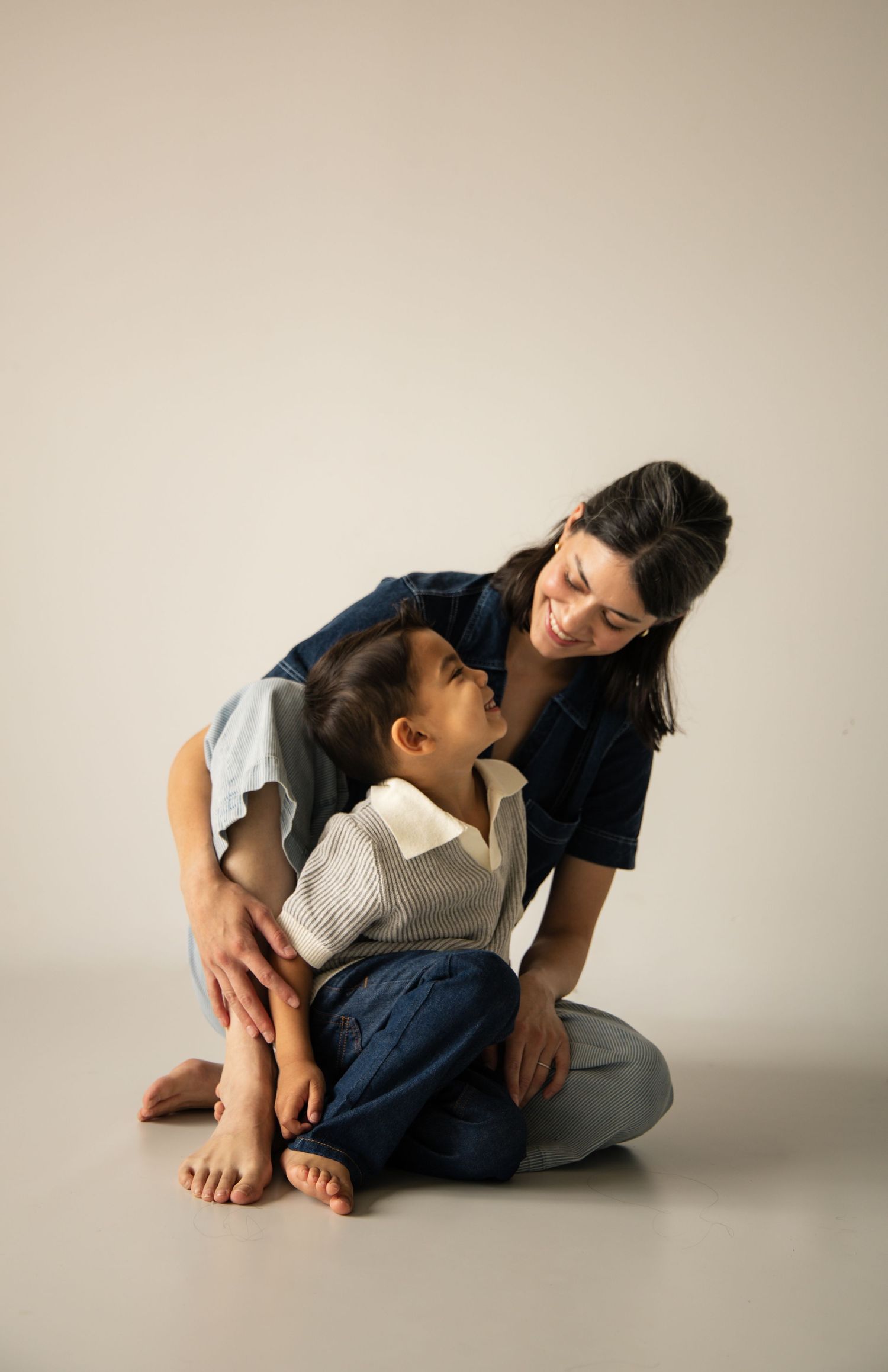 A smiling woman sits on the floor hugging a young child who looks up at her lovingly in a studio setting.