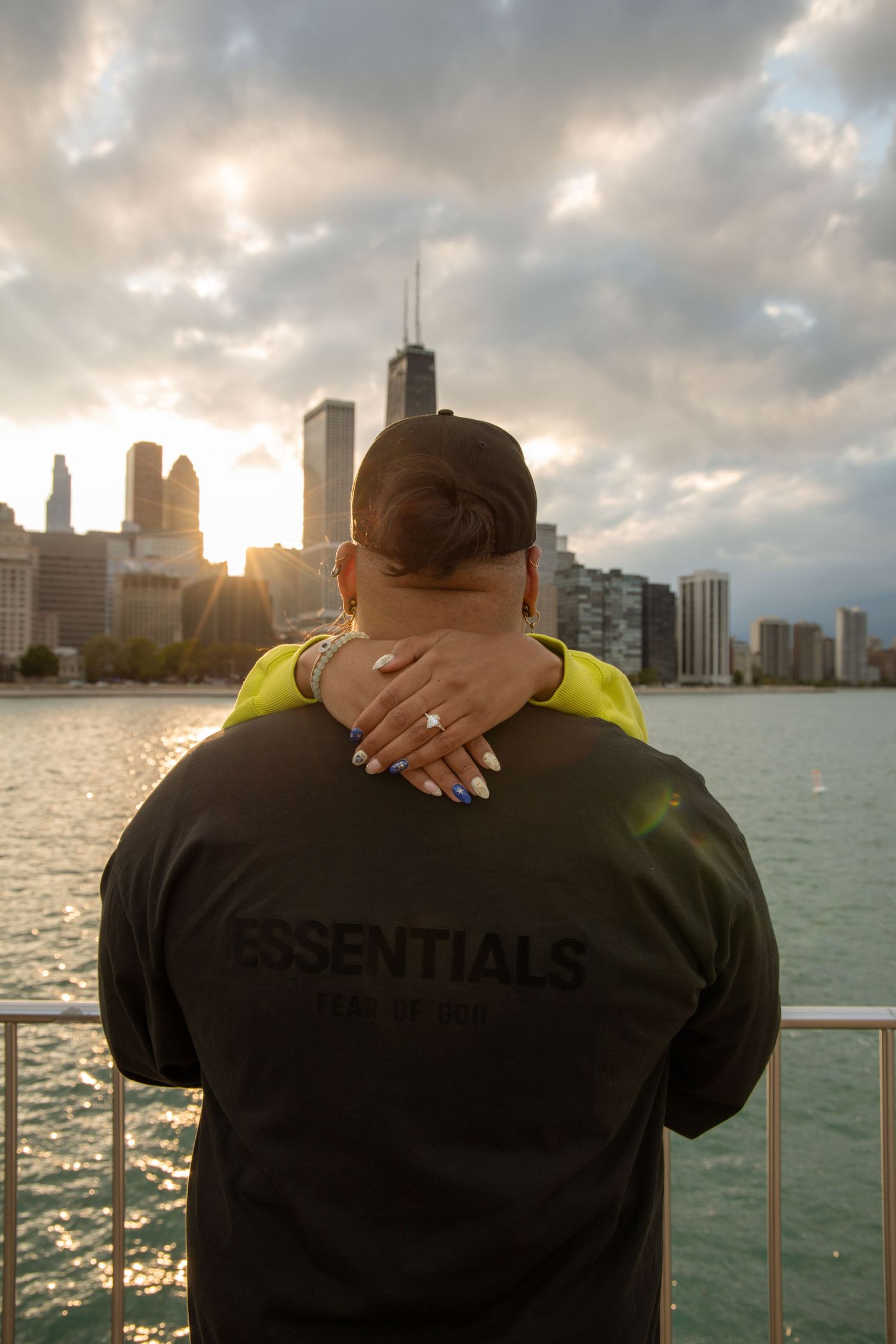 Couple embracing at Chicago waterfront sunset, man wearing Essentials Fear of God hoodie with city skyline backdrop.