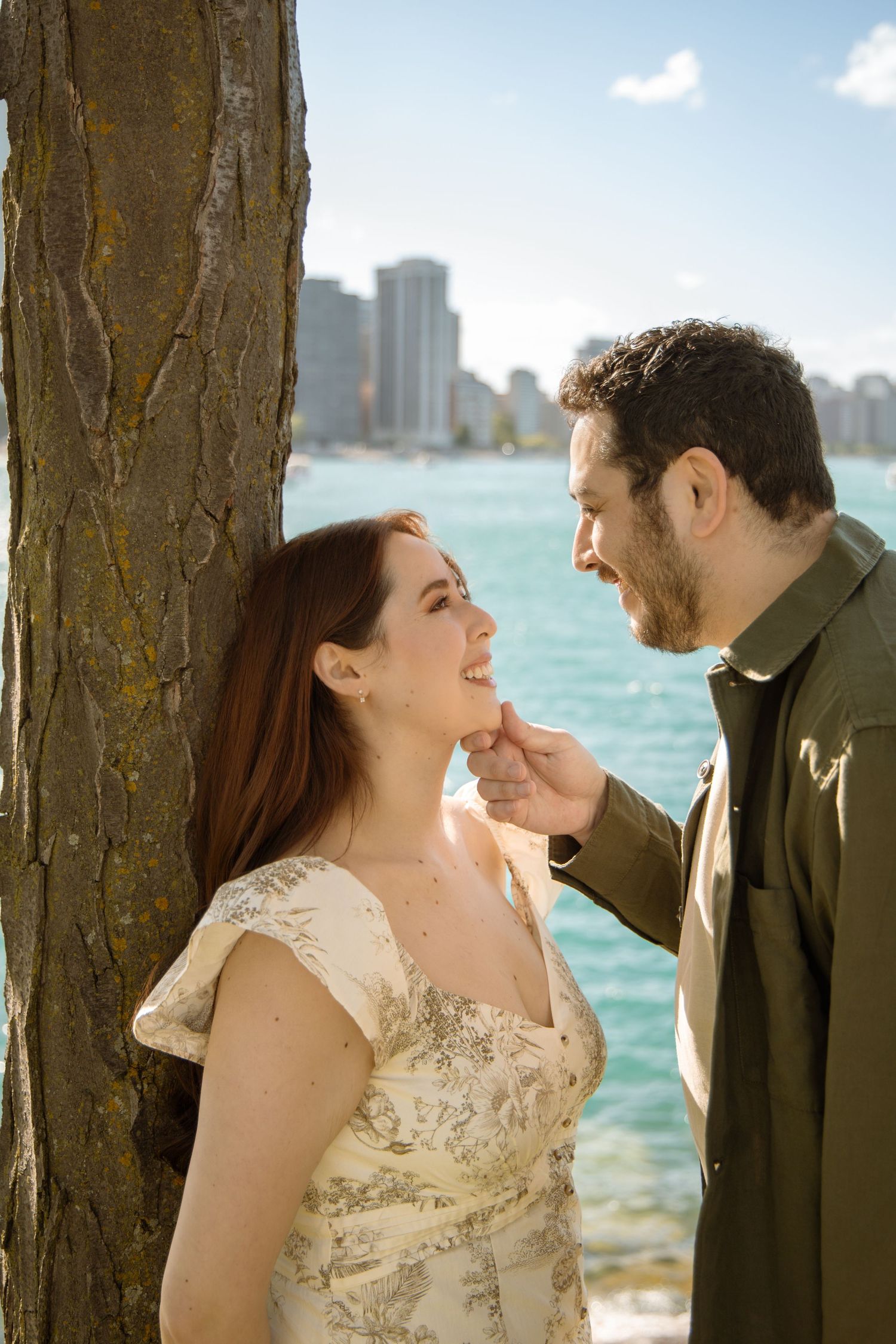 Couple sharing a romantic moment by a tree near the waterfront with a city skyline in the background.