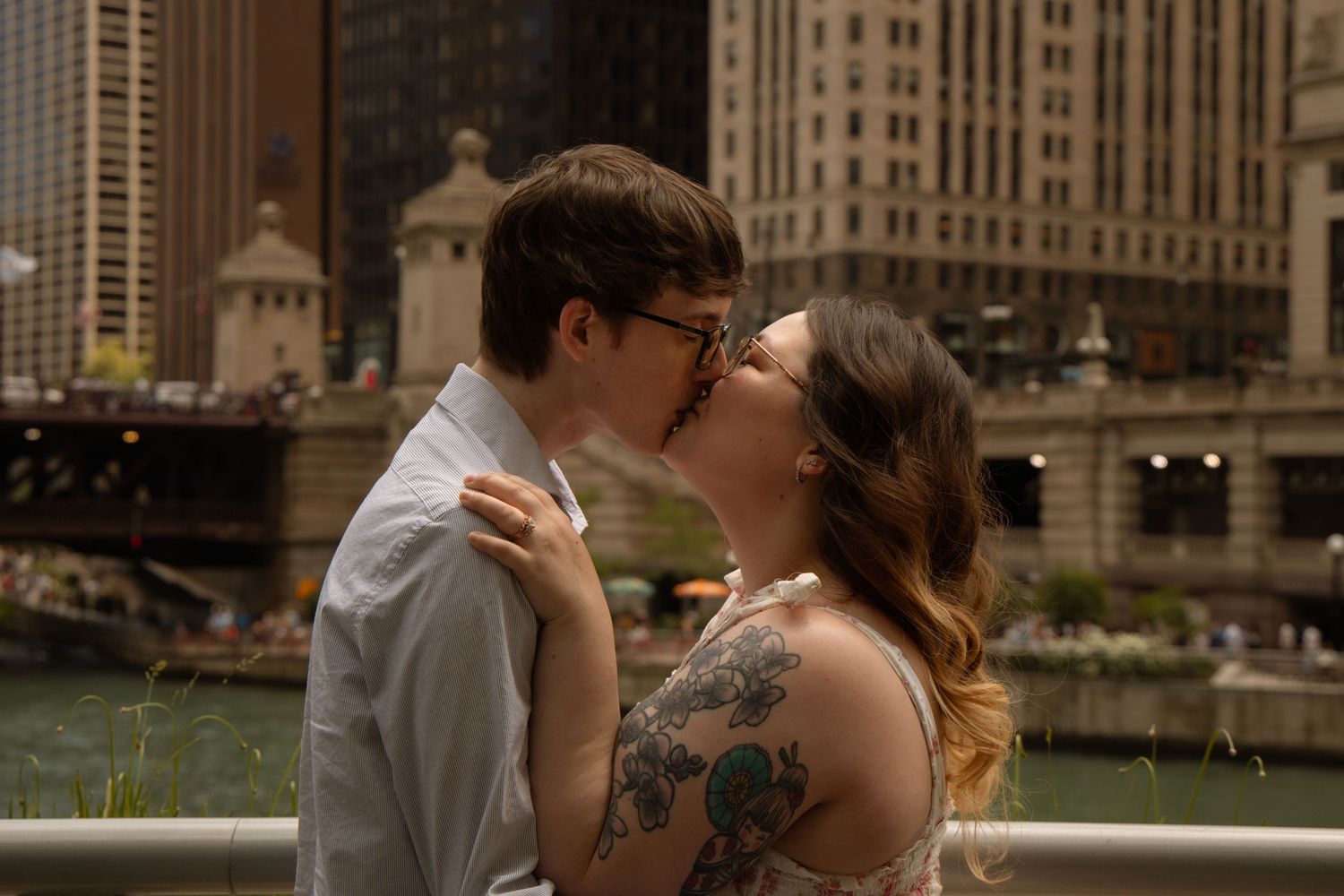 Couple sharing a romantic kiss along the Chicago River with city skyline and bridges in the background.