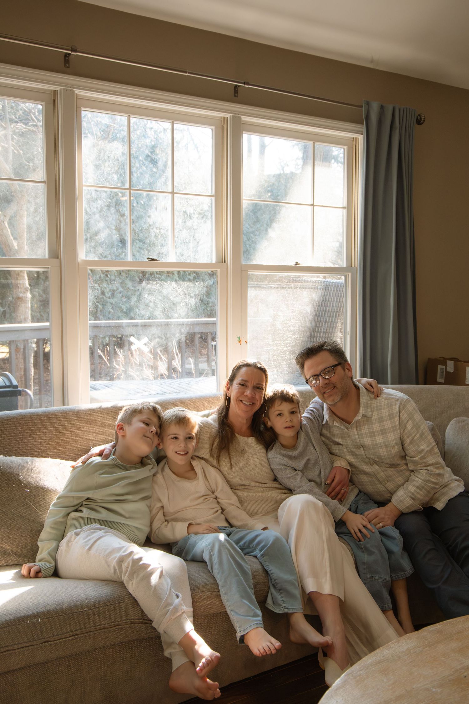 A happy family of five sitting together on a couch in a cozy living room, smiling in front of bright windows.