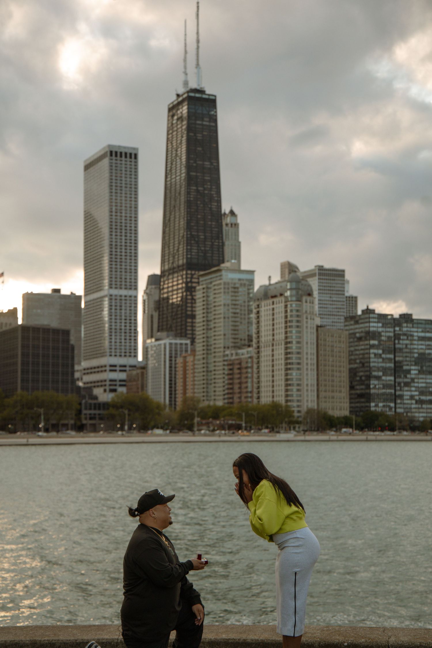 Man proposes to woman on one knee by Chicago waterfront with iconic John Hancock Center skyline backdrop.
