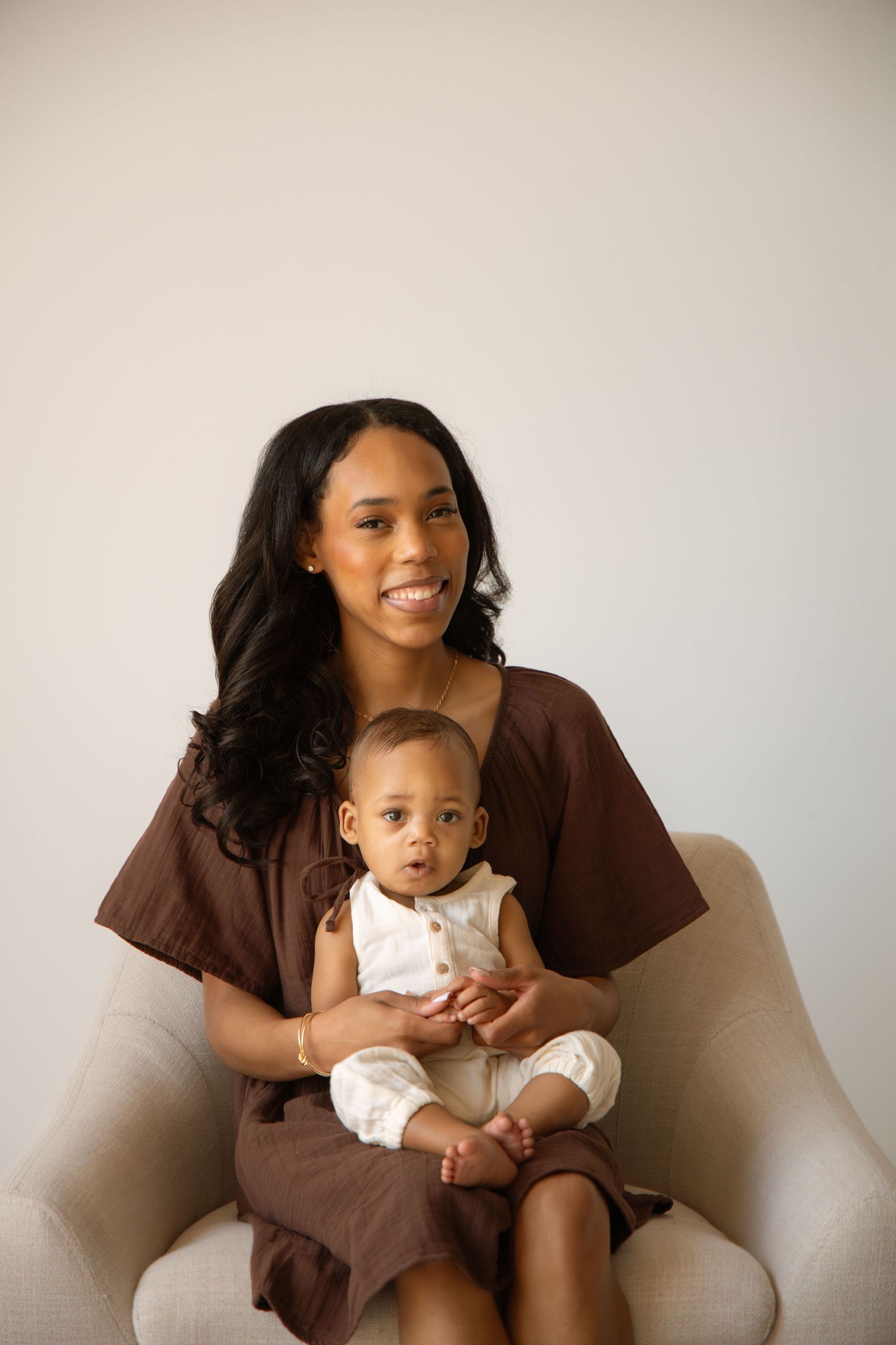 A smiling woman in a brown dress sits in a chair holding a baby dressed in white.