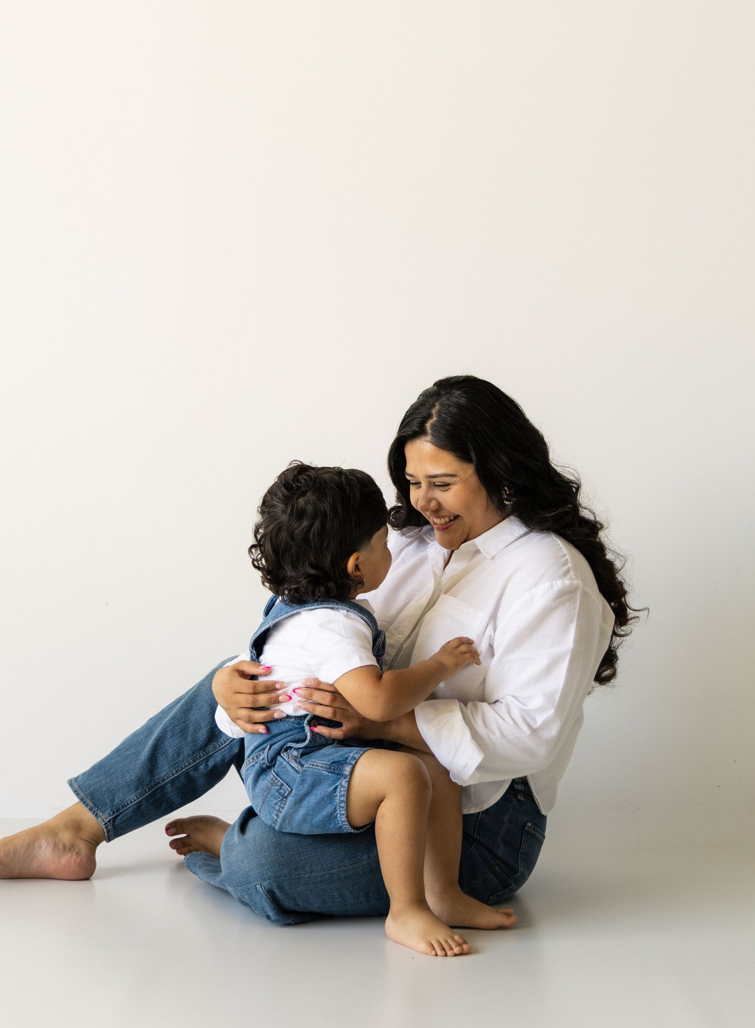A mother sits on a white studio floor smiling at her toddler in denim overalls, sharing a tender moment.