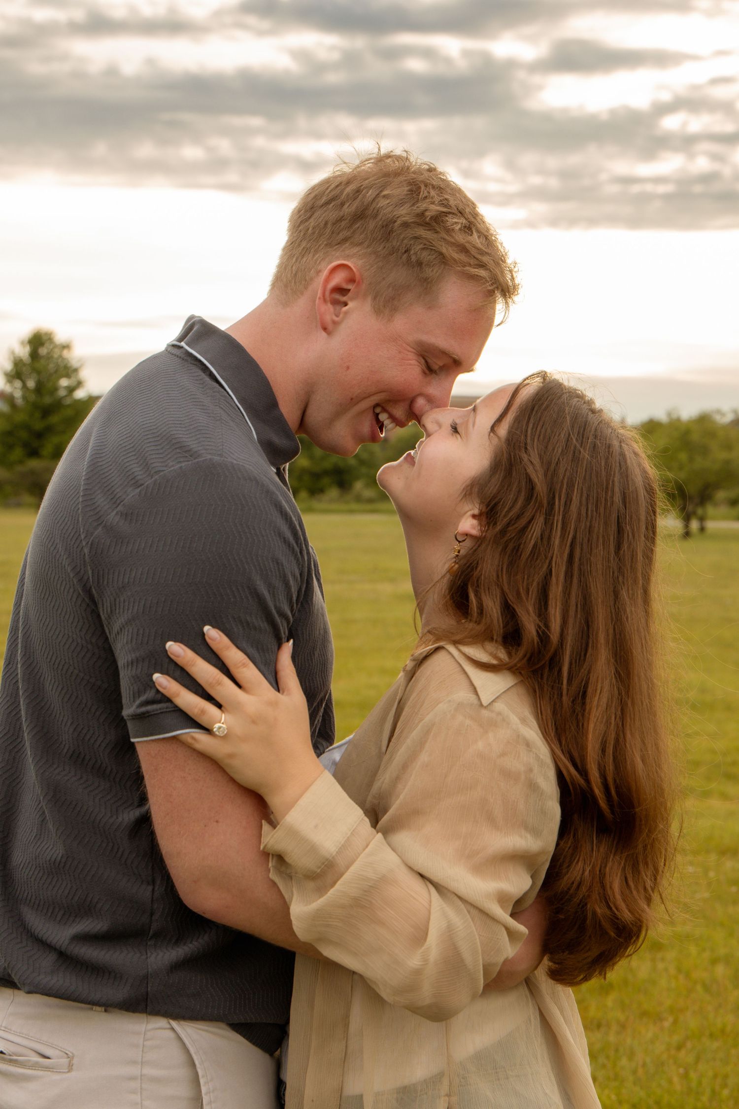 A smiling couple embraces and nearly kisses in a green field at sunset, the woman wearing an engagement ring.