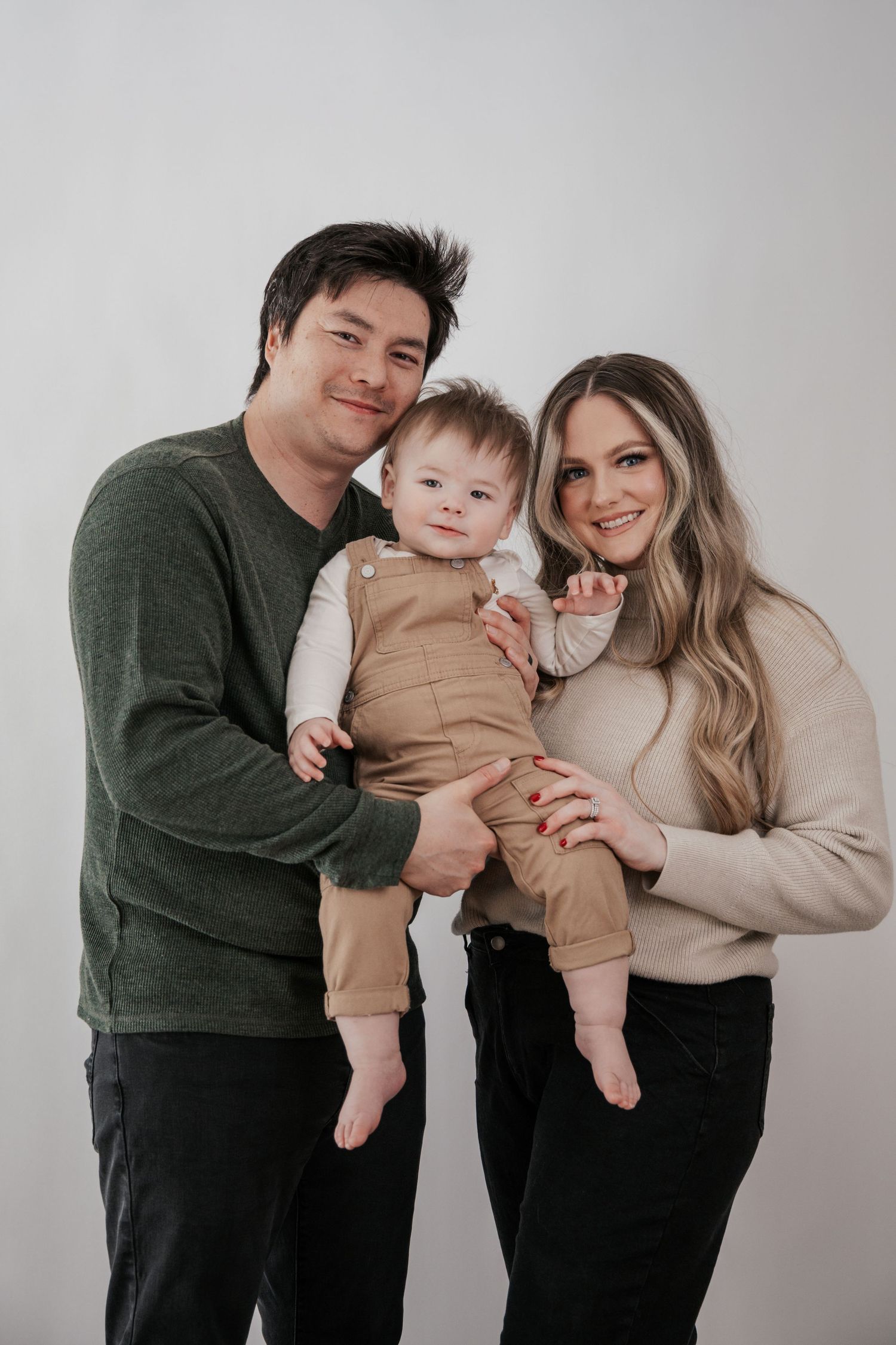 Smiling parents in a studio portrait hold their baby dressed in brown overalls and a white shirt.