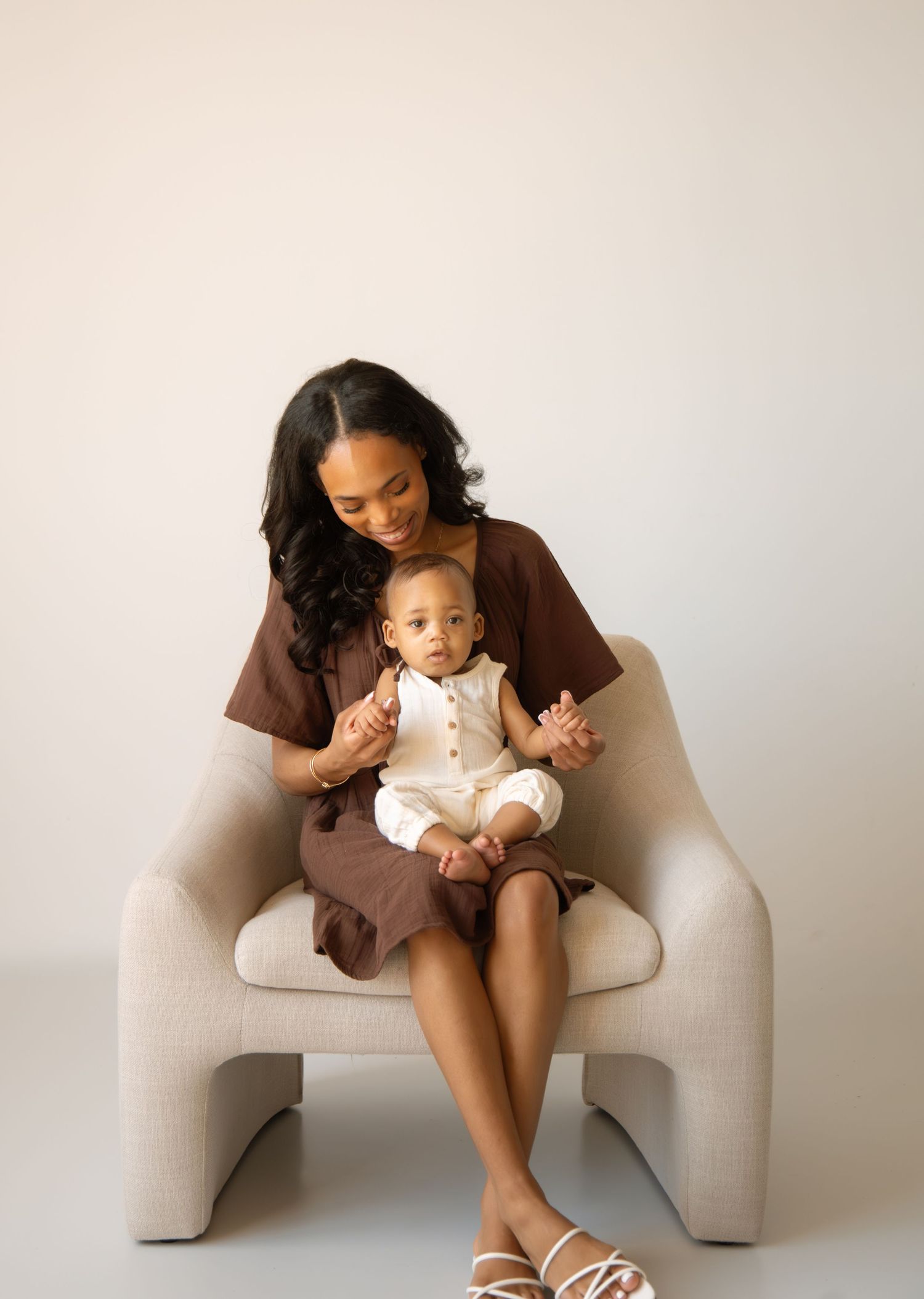 A mother in a brown dress sits in a cream chair, lovingly holding her smiling baby in a white outfit.