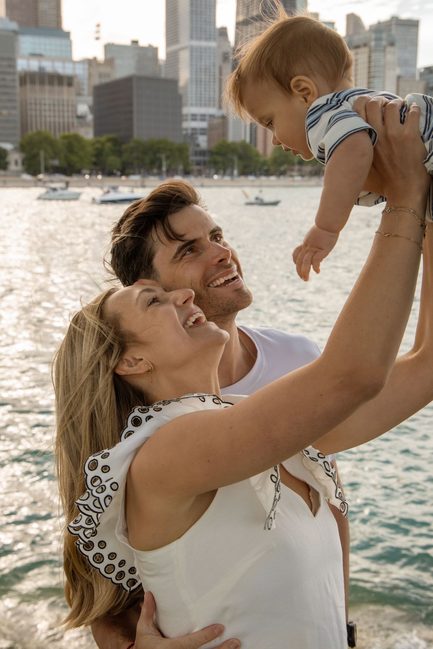 Happy young family with baby by Chicago waterfront, parents lifting smiling infant against city skyline backdrop.