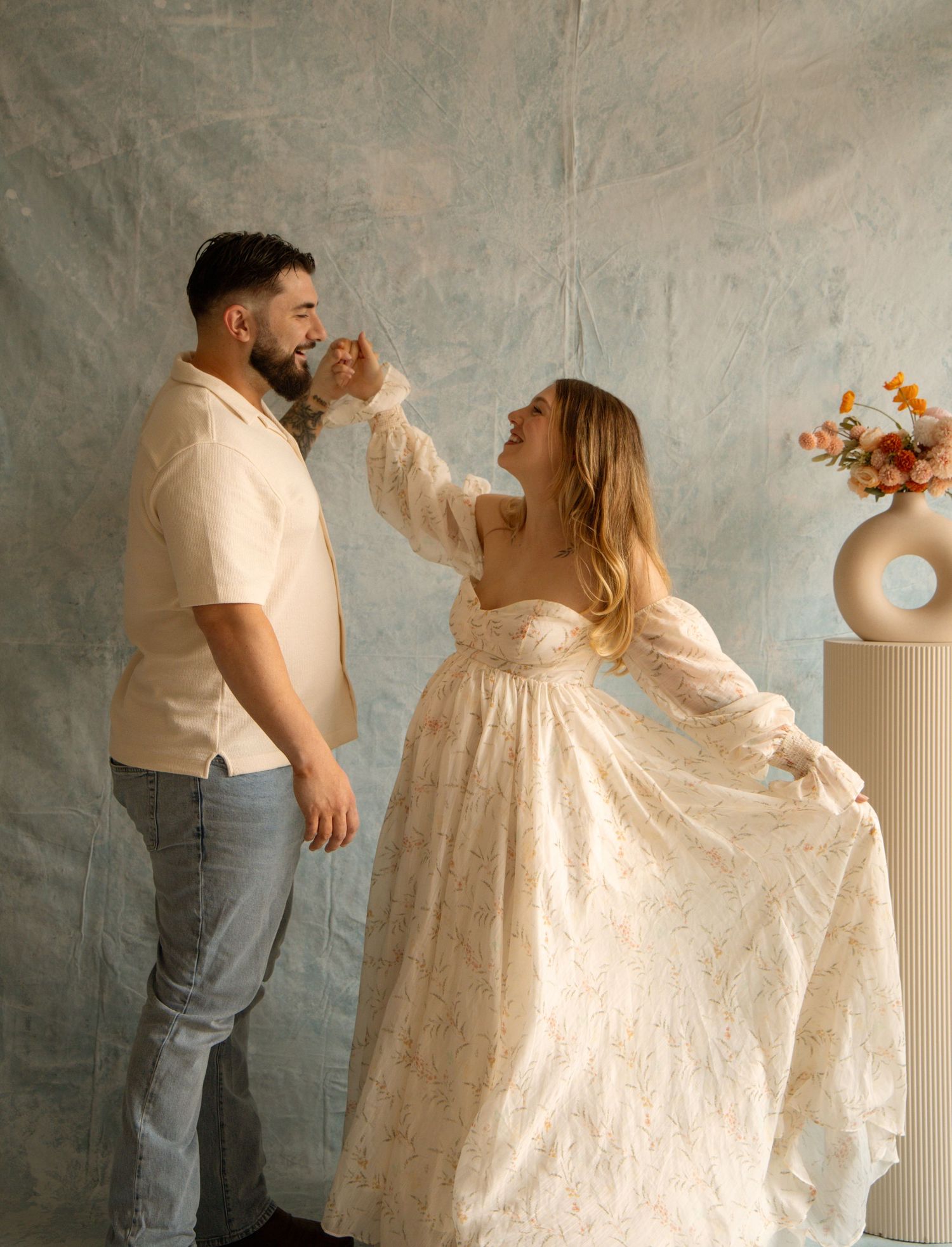 Couple dancing together in a studio, woman in floral gown holding man's face, floral arrangement in background.