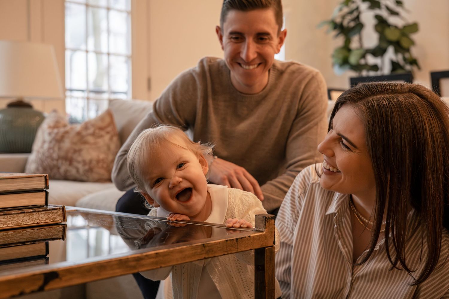 Happy family with parents smiling at laughing baby standing at wooden coffee table in cozy living room.