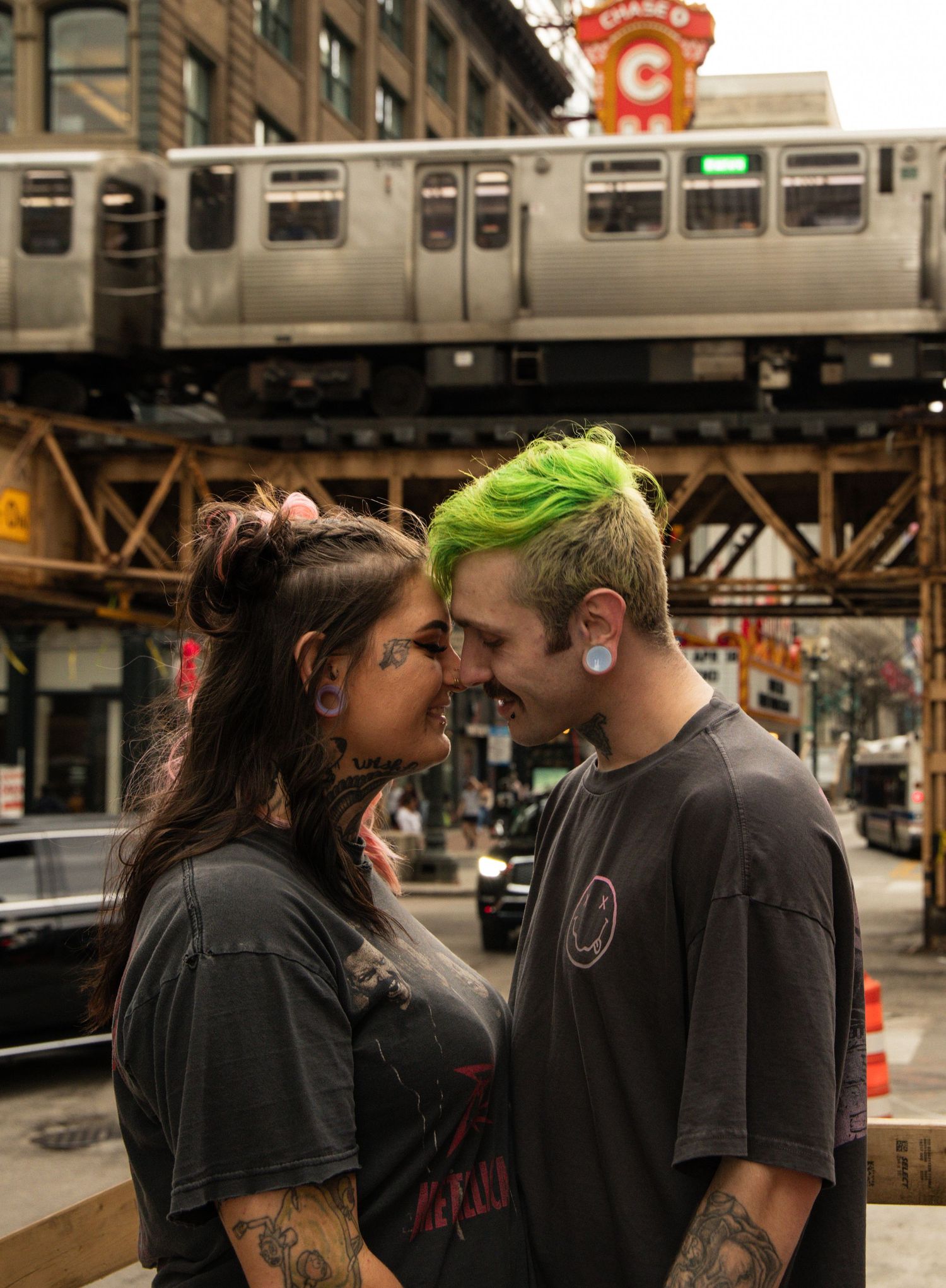 A couple with tattoos and green hair share a romantic moment on a Chicago street with the elevated train above.