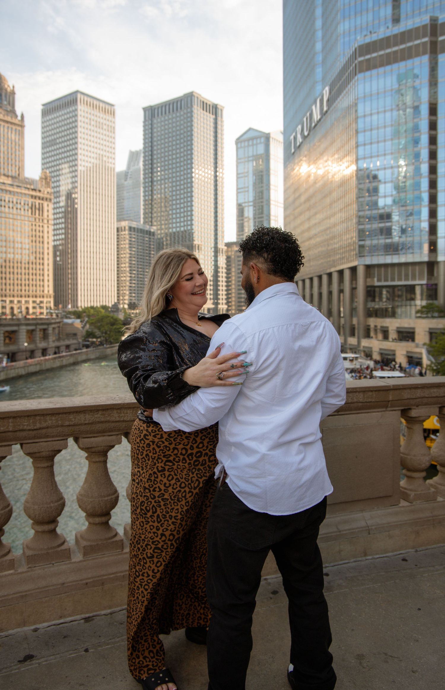Happy couple embraces on Chicago Riverwalk with Trump Tower and city skyline behind them.