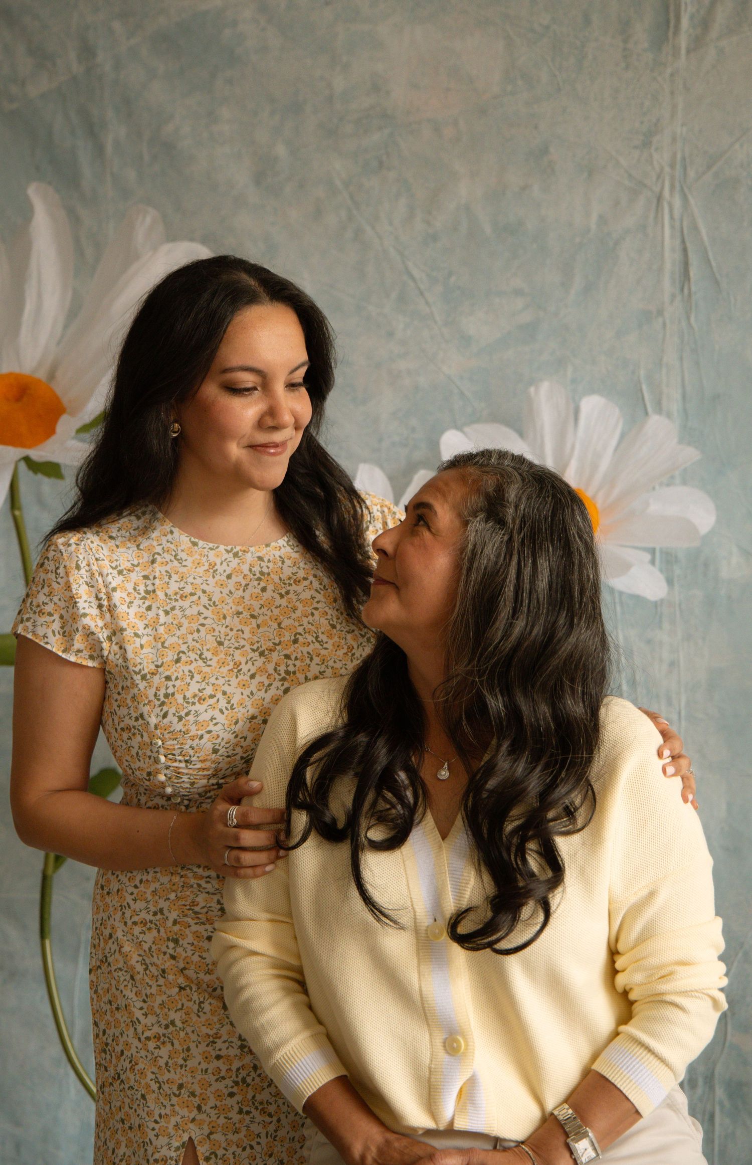 Two women sharing a warm moment together in front of a floral backdrop, one in a floral dress and one in a cream cardigan.
