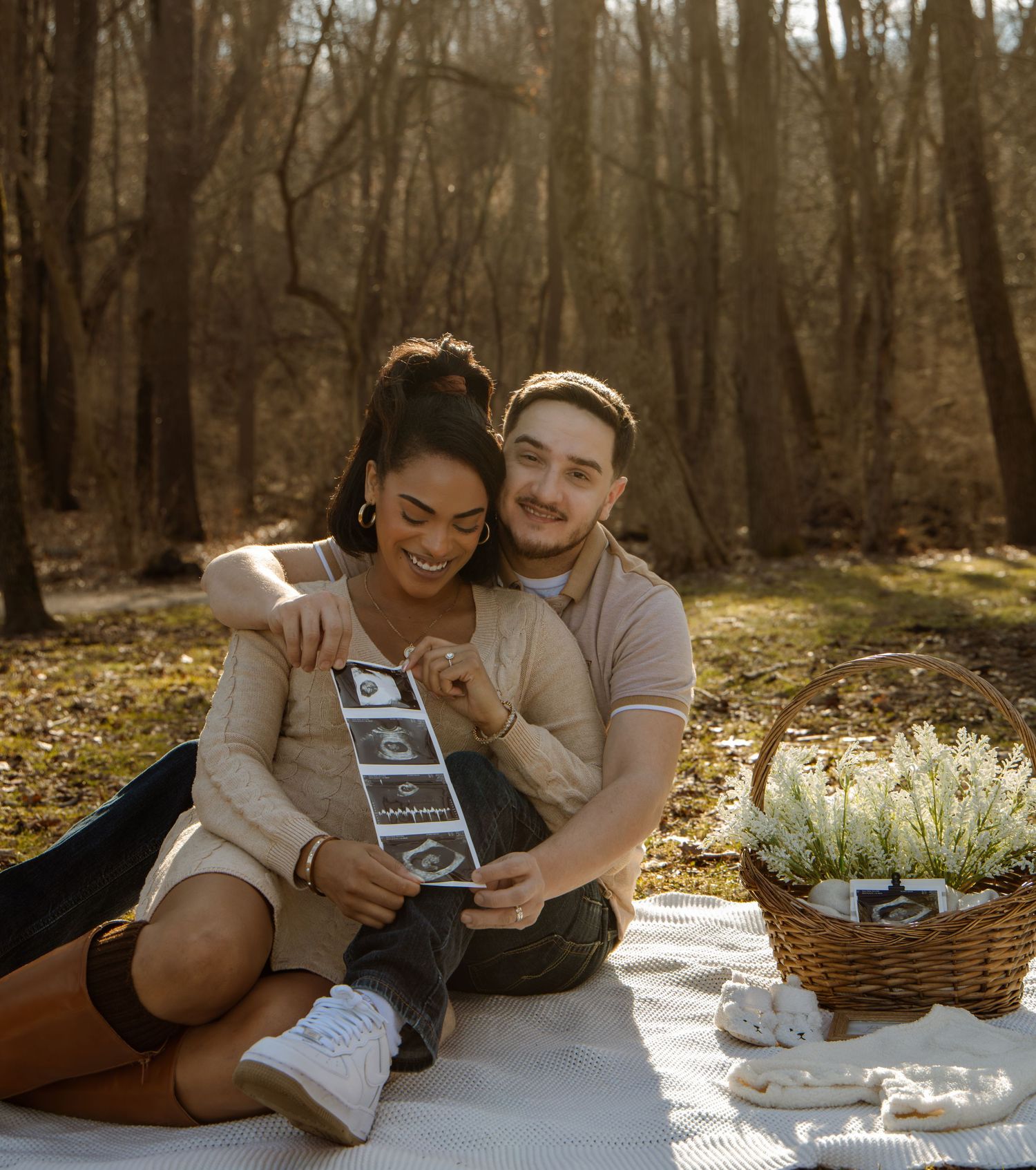 Smiling couple sitting on a blanket outdoors holding ultrasound photos with a flower basket nearby.