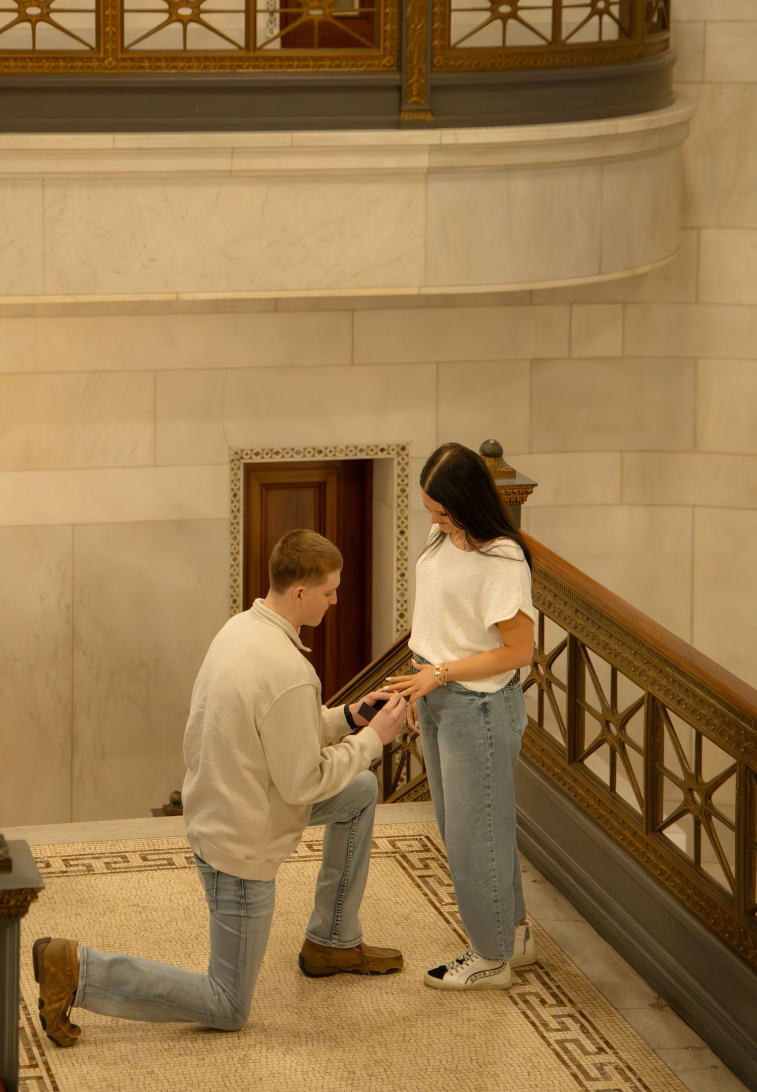 Man on one knee proposing to a woman on a staircase inside an ornate building.