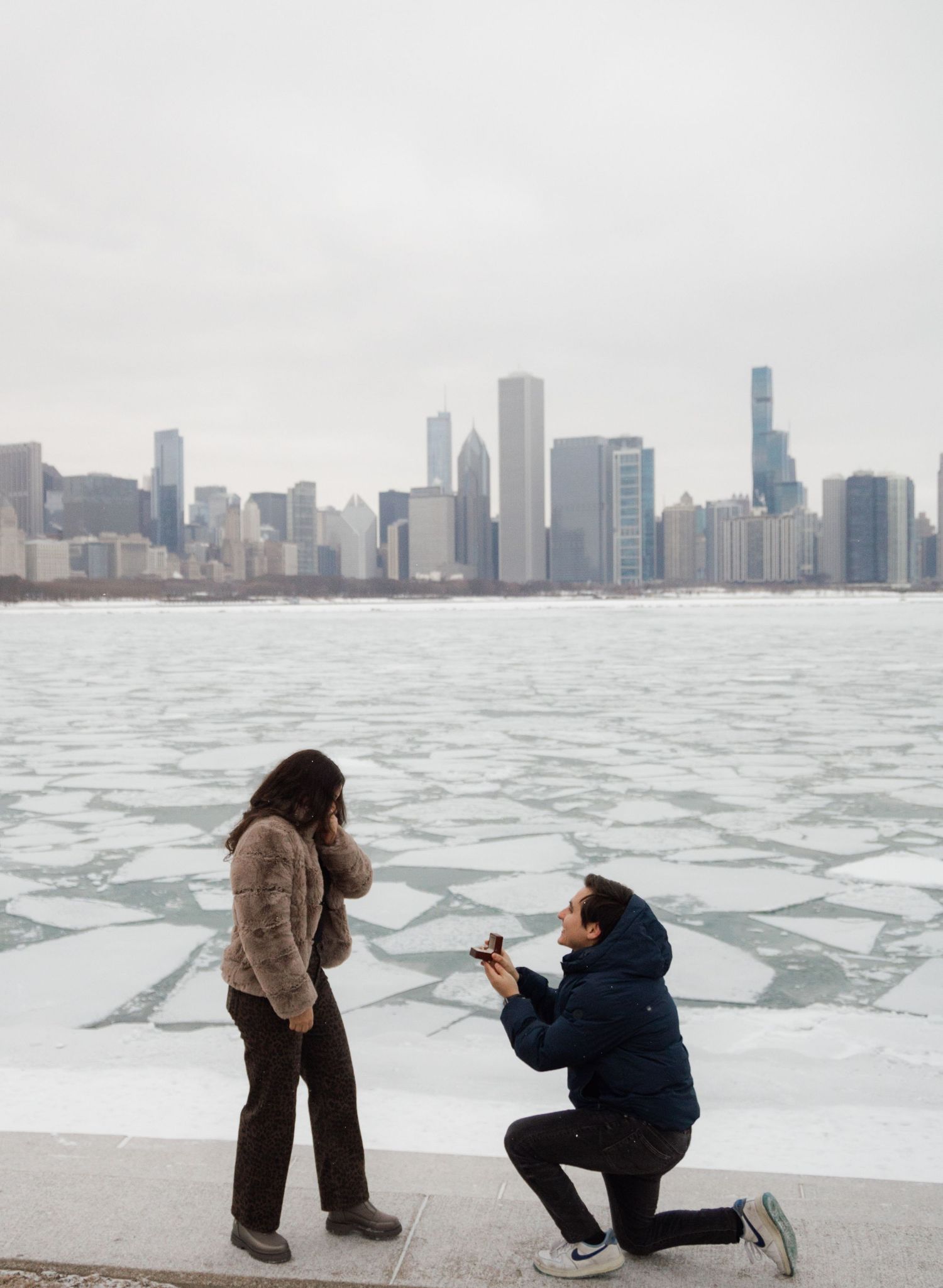 Man proposes to woman on Chicago lakefront with icy water and city skyline behind them.