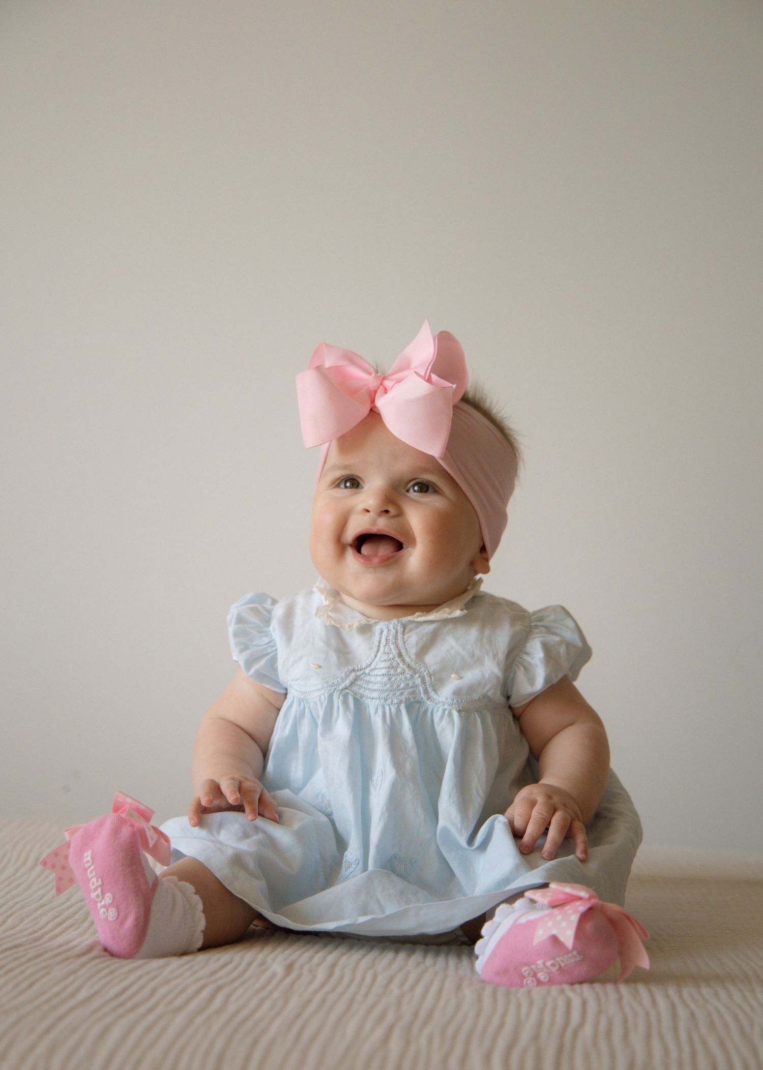Smiling baby girl in light blue dress with pink bow headband and pink socks sitting on white surface.
