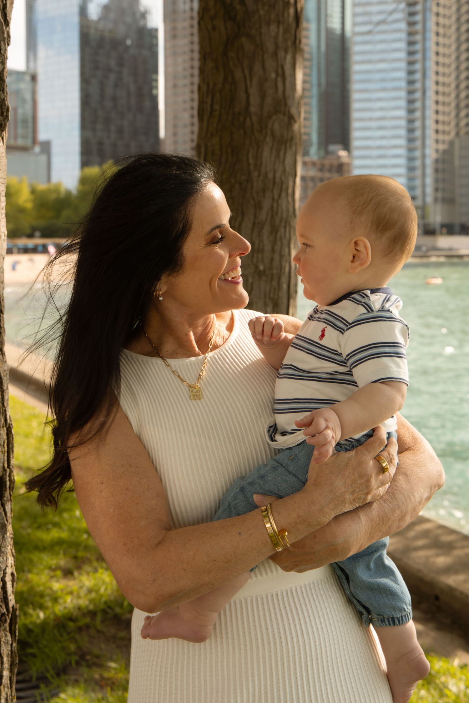 A smiling woman in a white dress holds a happy baby boy near a tree with a city skyline and waterfront in the background.