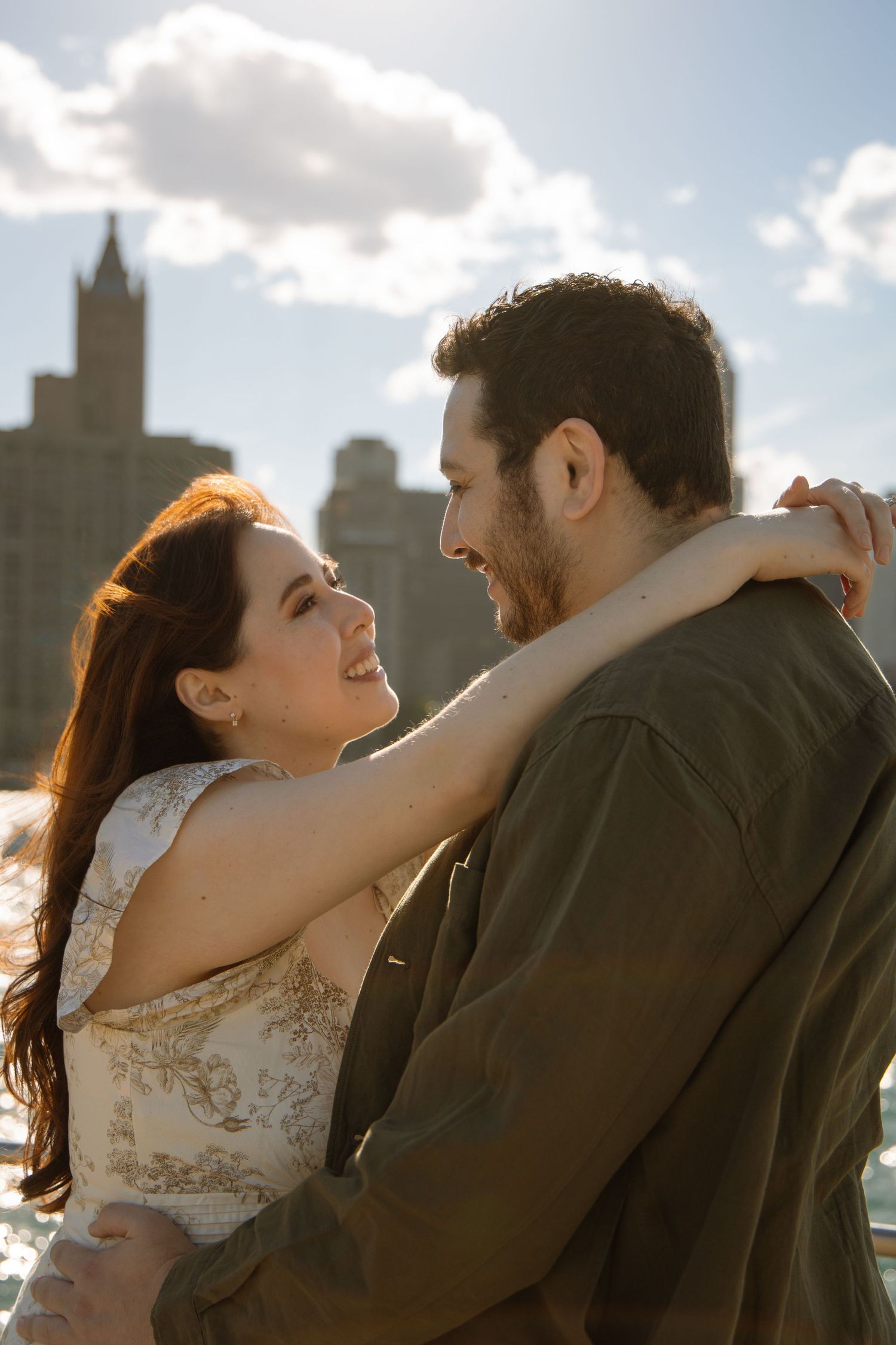 A couple embraces and smiles at each other with a city skyline and cloudy sky in the background.