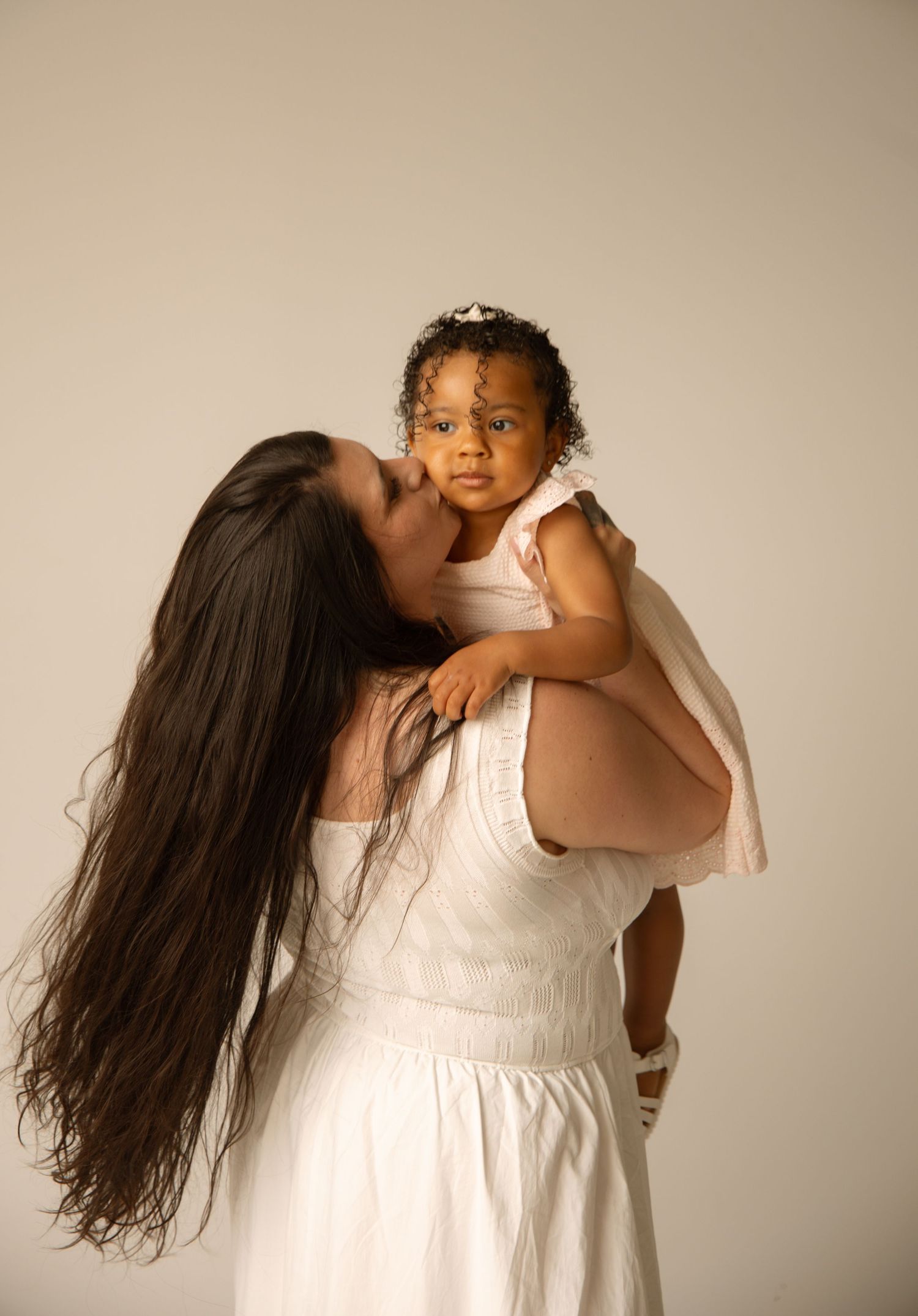 A mother in a white dress with long dark hair kisses her baby girl in a studio portrait.