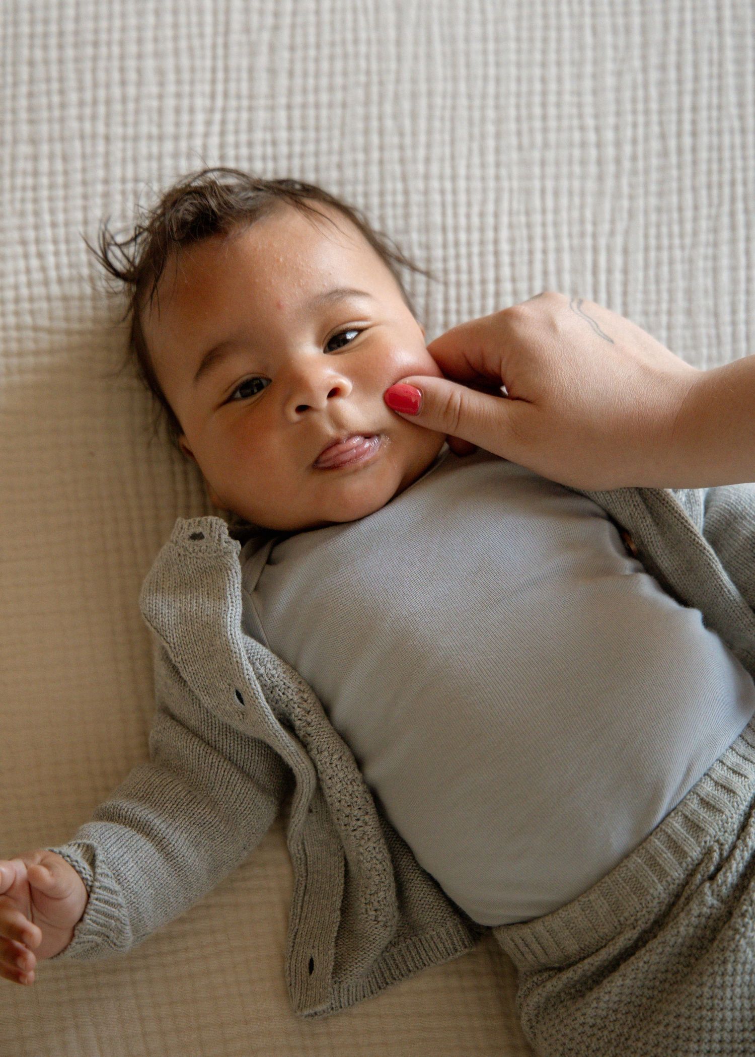 A cute baby in a gray outfit lying on a white blanket while an adult gently touches their chin.