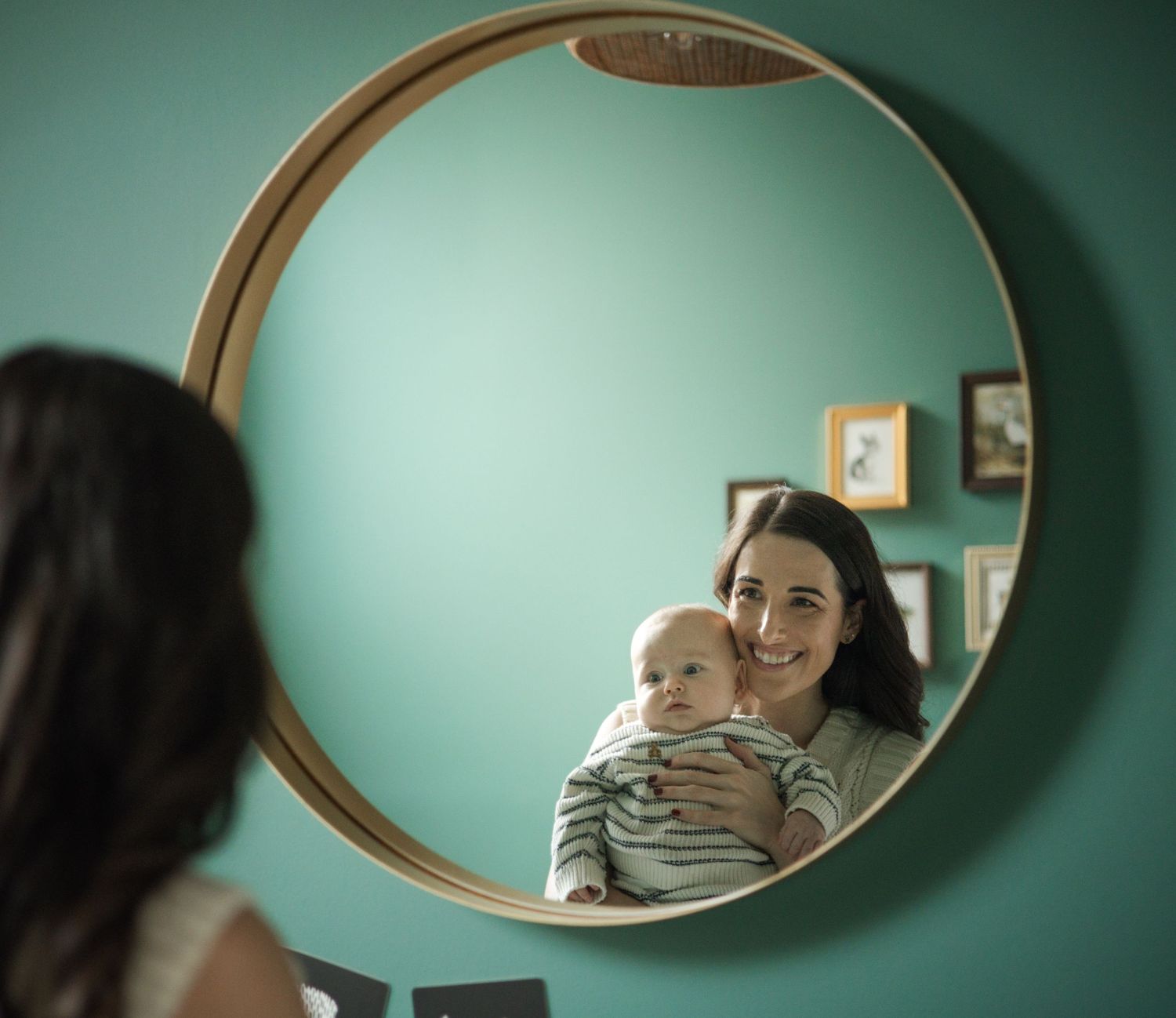 A smiling mother holding her baby reflected in a round gold-framed mirror on a teal wall.