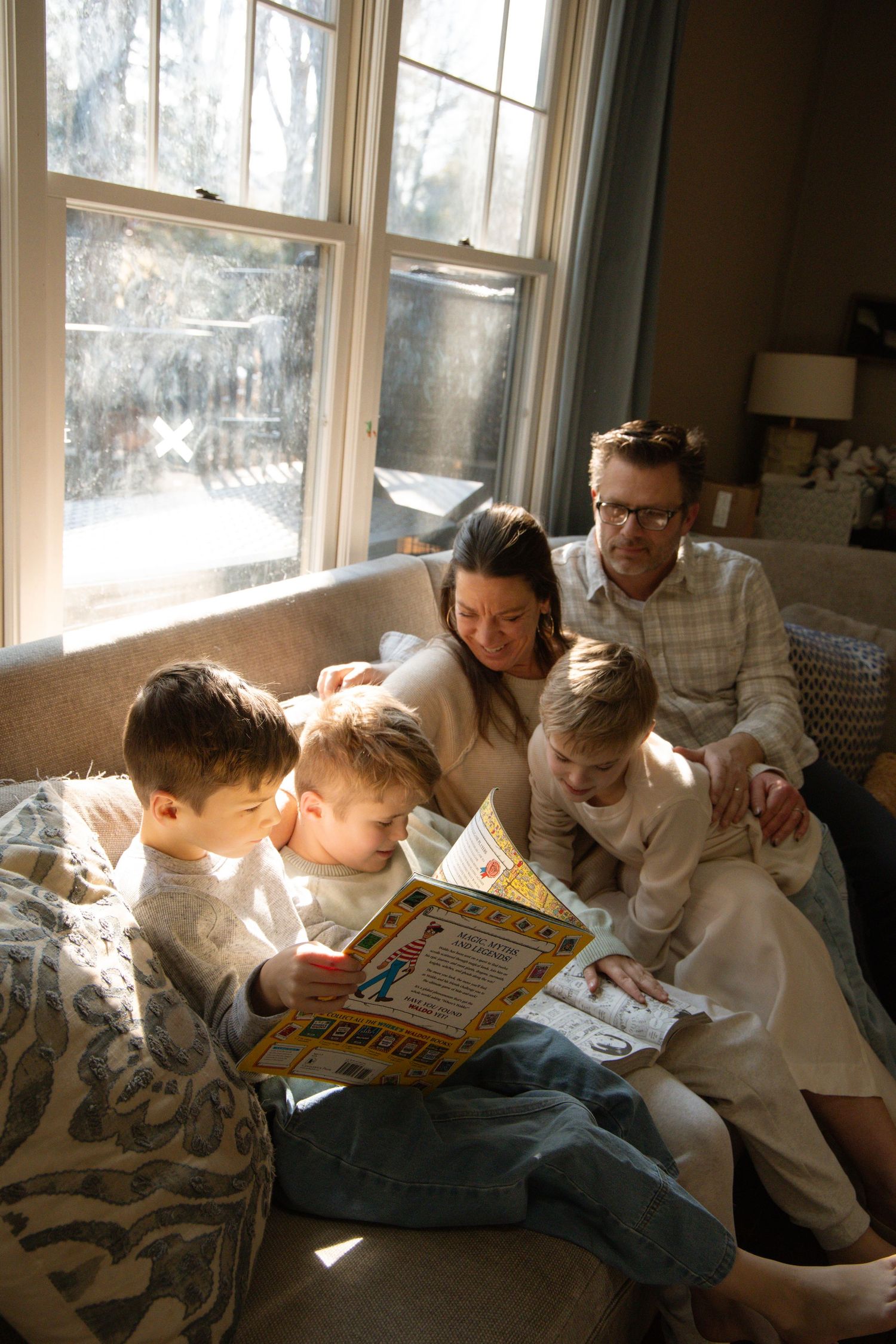 Family of five reading a colorful children's book together on a cozy sunlit sofa near a large window.