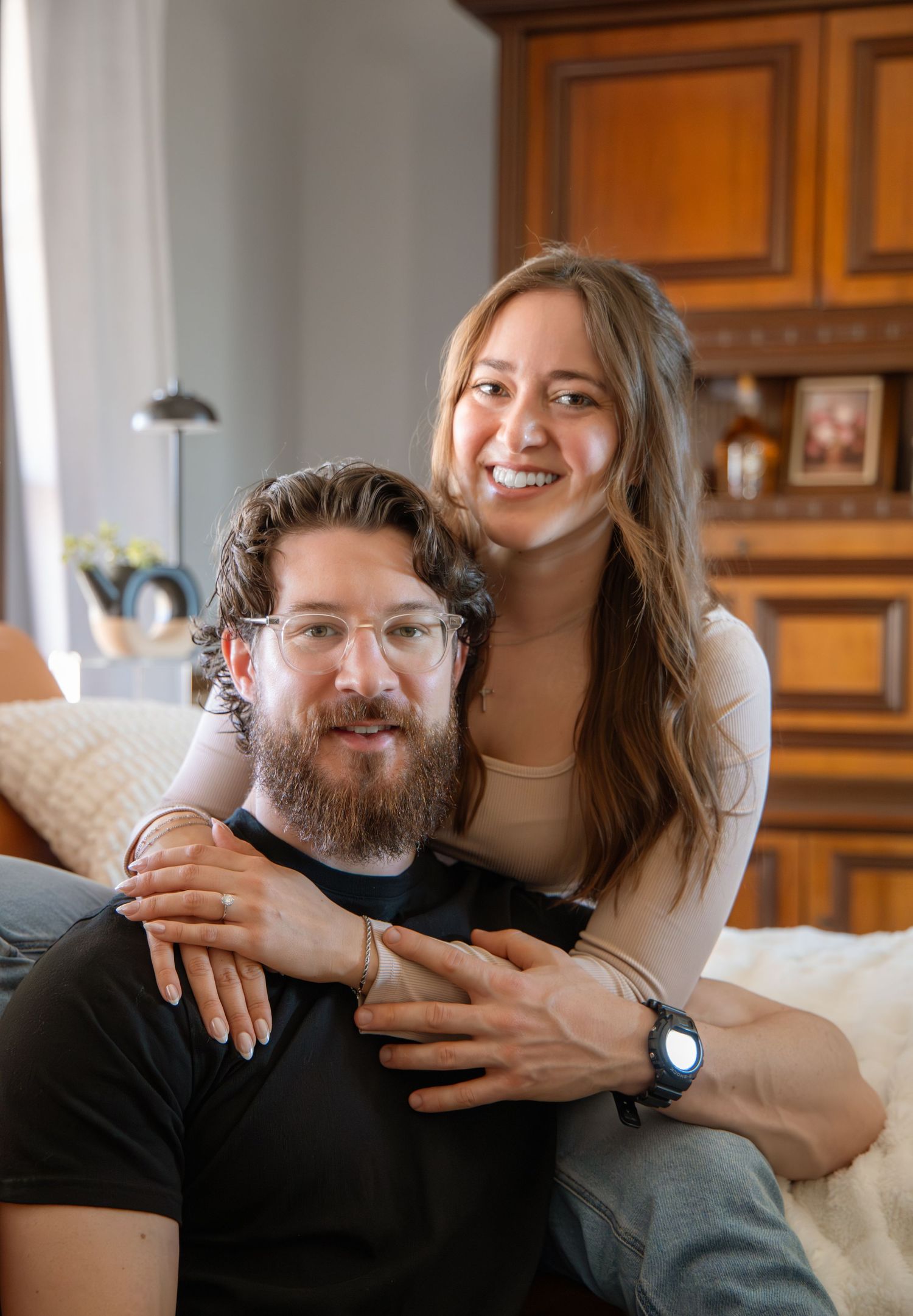 A smiling couple poses together indoors, man with beard and glasses in black shirt, woman in beige top hugging him from behind.