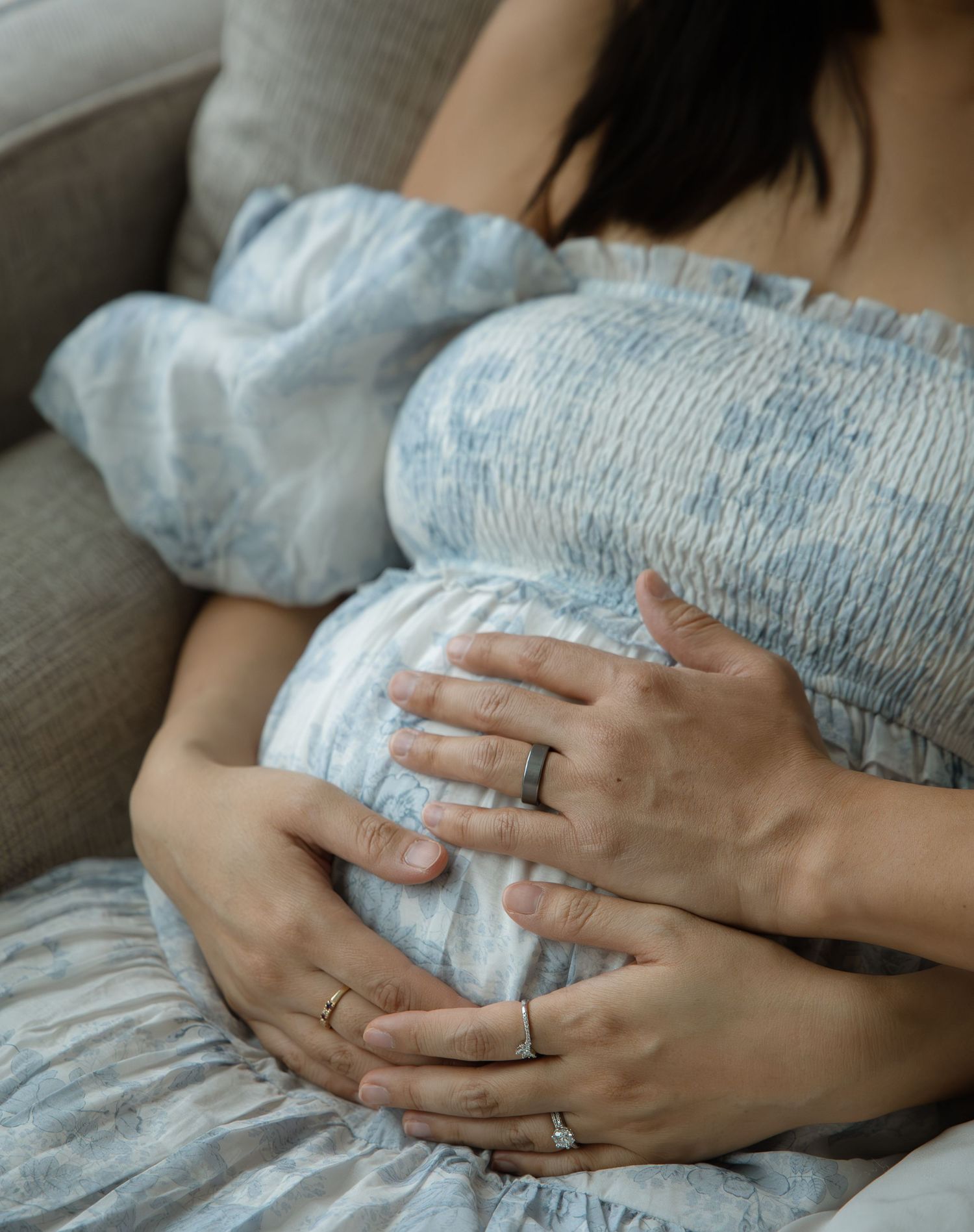 Pregnant woman in blue floral dress cradling baby bump while sitting on couch, wearing gold and silver rings.