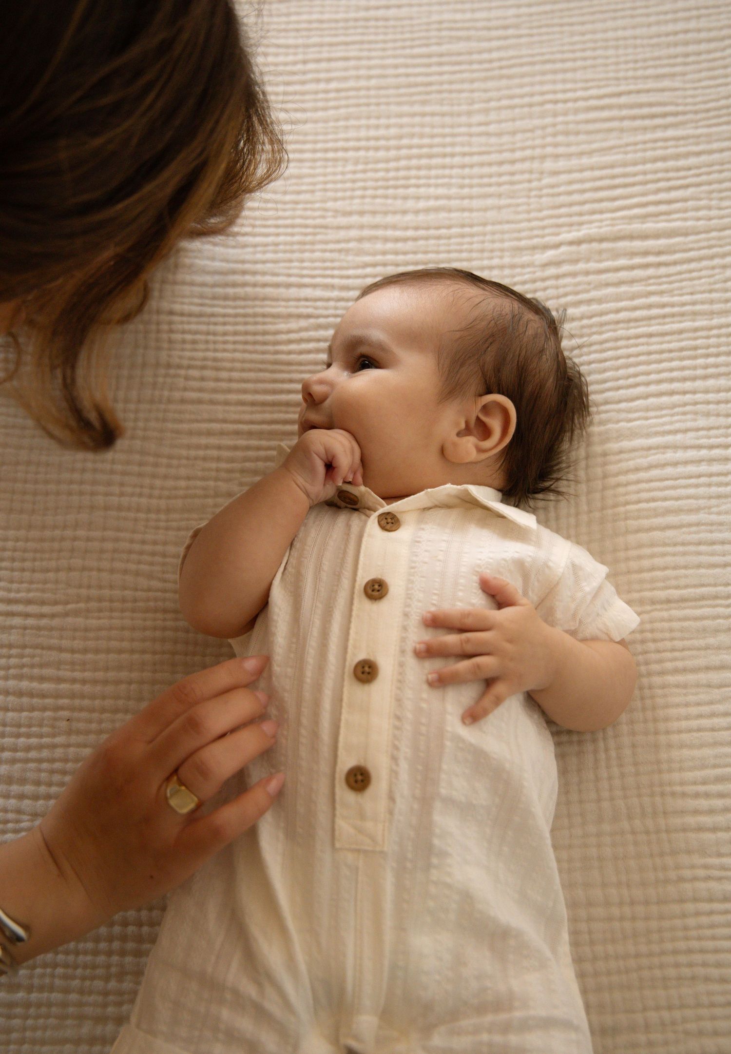 A baby in a cream linen romper lies on a white blanket while a mother lovingly gazes down at them.