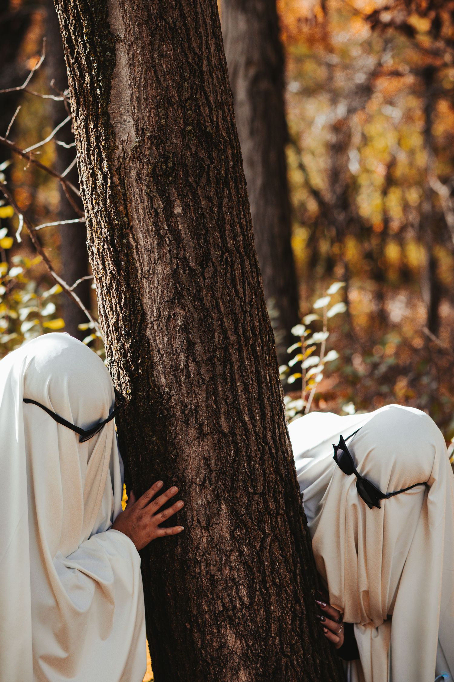 Two people in white ghost costumes peek around a tree trunk in an autumn forest.