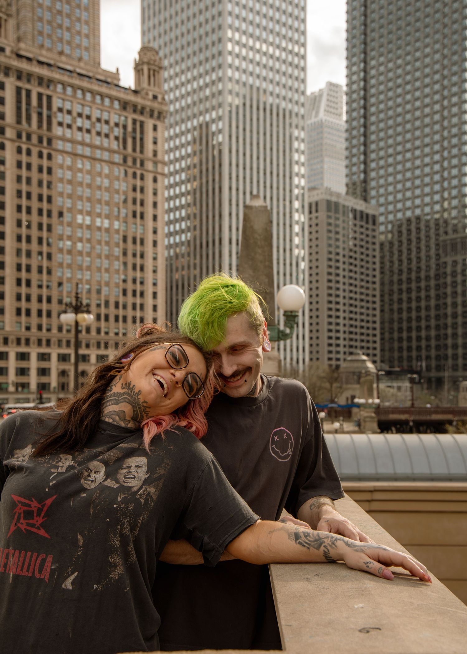 Two tattooed people with colorful hair laugh together on a rooftop with Chicago skyscrapers in the background.