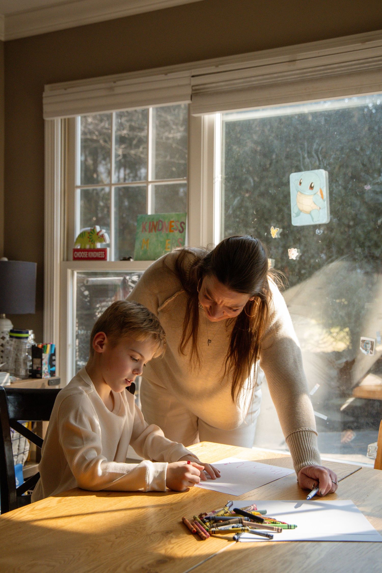 A woman helps a young child with drawing at a wooden table, with crayons nearby and sunlight streaming through windows.