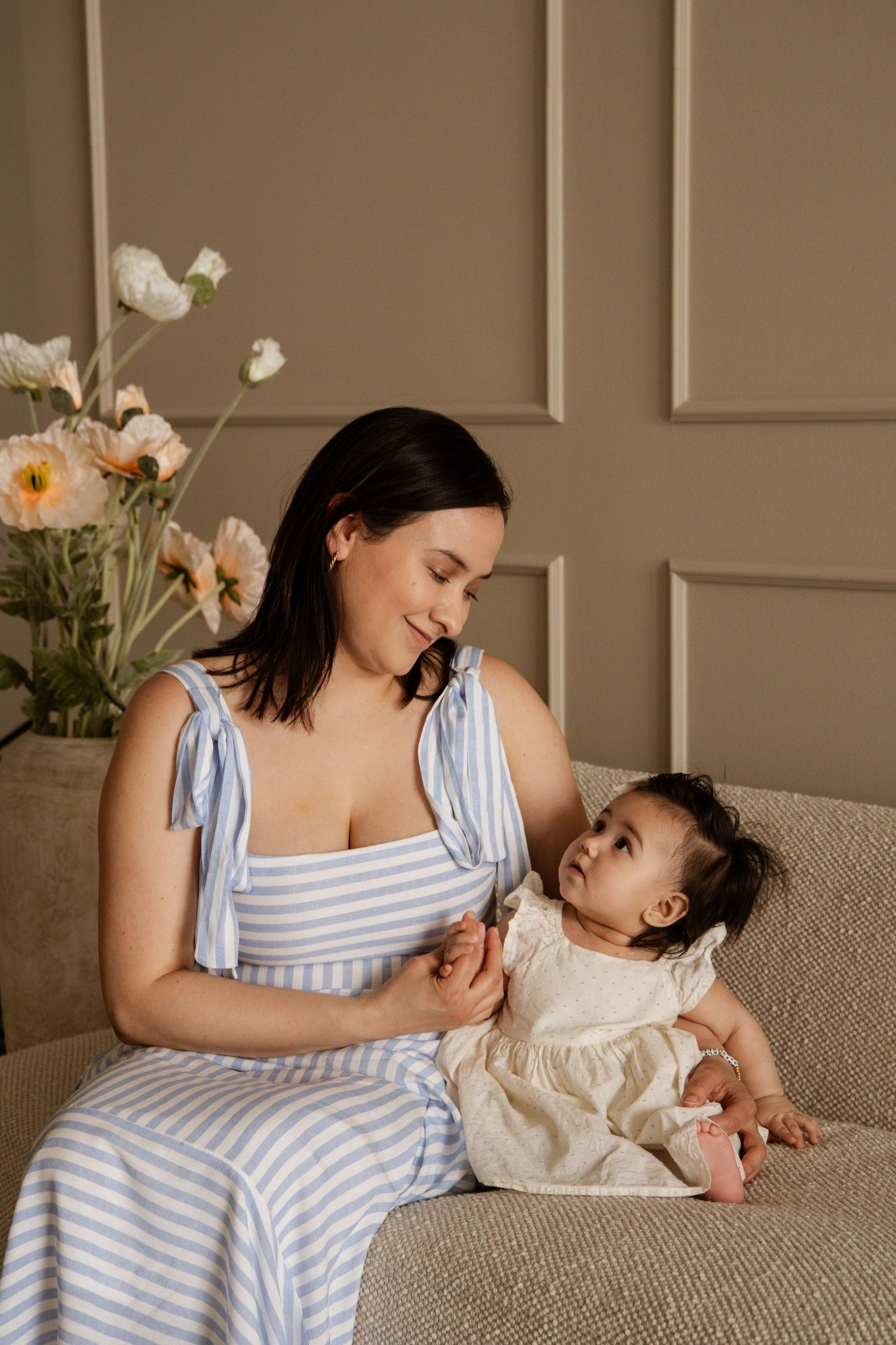 A smiling mother in a blue striped dress sits on a sofa holding her baby girl in a white dress, with flowers in background.
