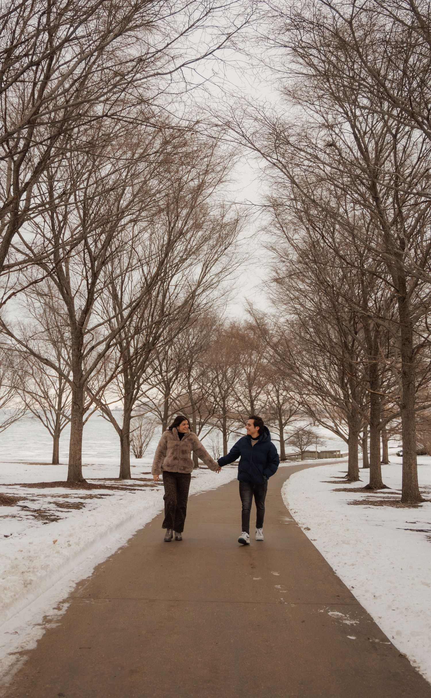 Couple holds hands walking down a snowy tree-lined path in a winter park.