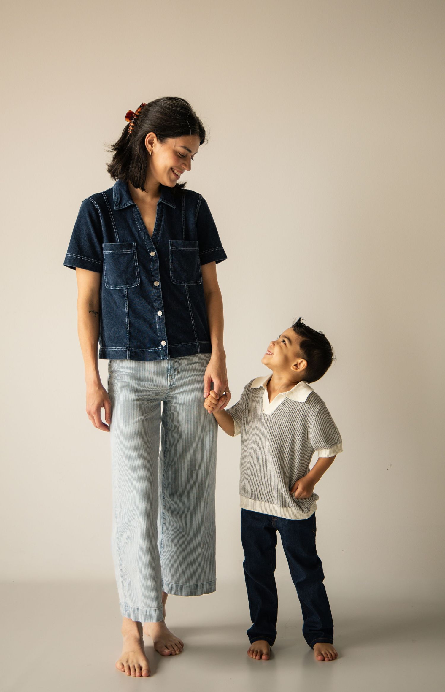 A woman in a denim shirt and light jeans holds hands with a young boy in a polo shirt, both smiling at each other.