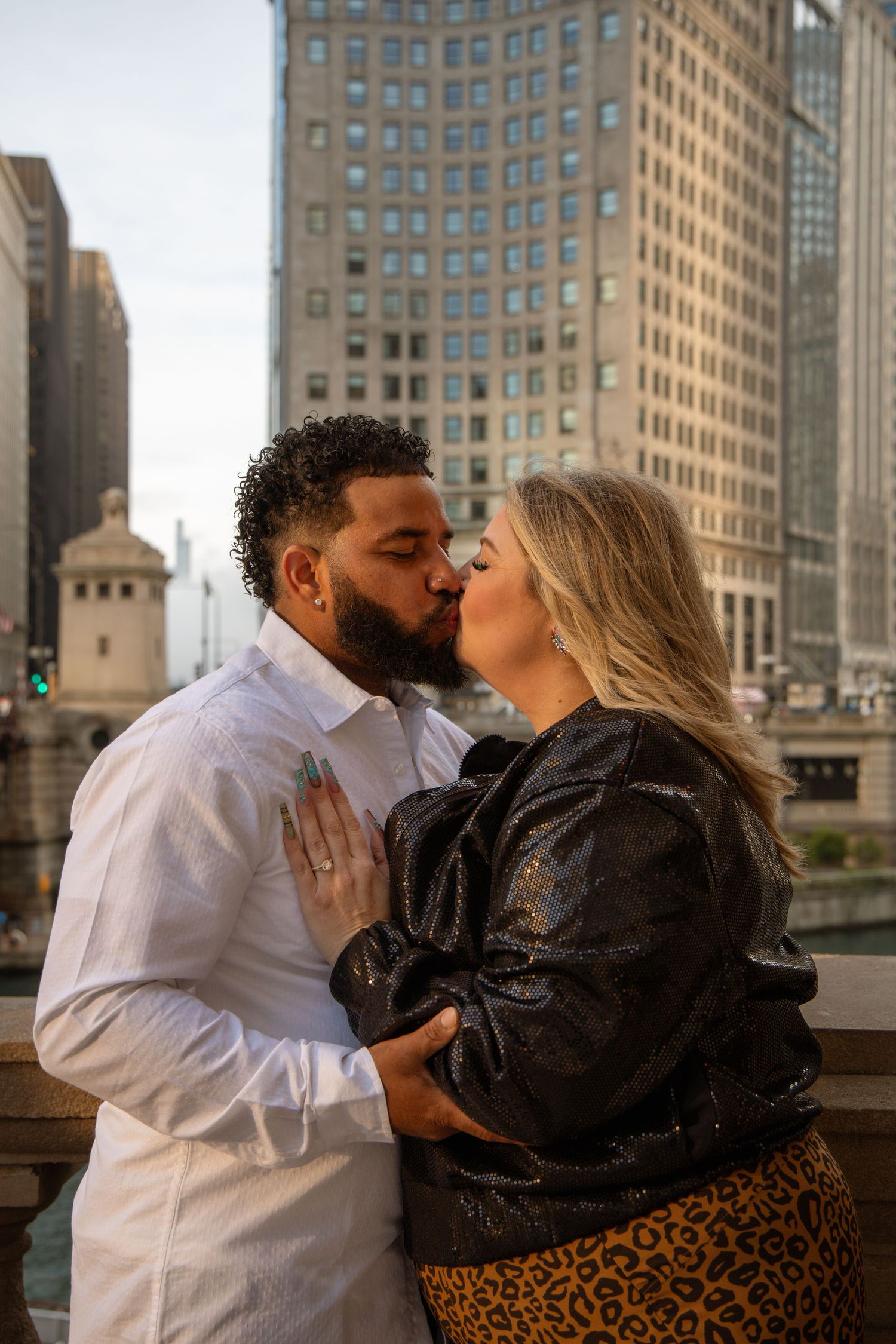 Couple sharing a romantic kiss on a Chicago riverwalk bridge, with city skyscrapers in the background.