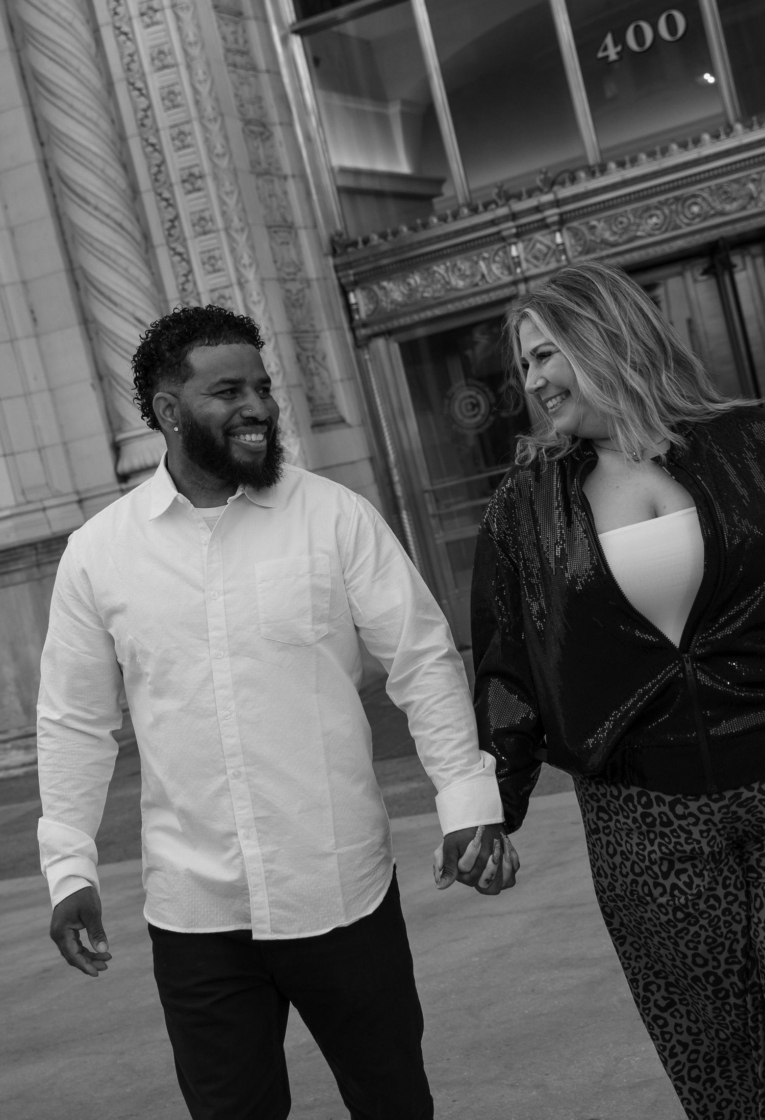 Black and white photo of a smiling couple holding hands outside a building numbered 400.