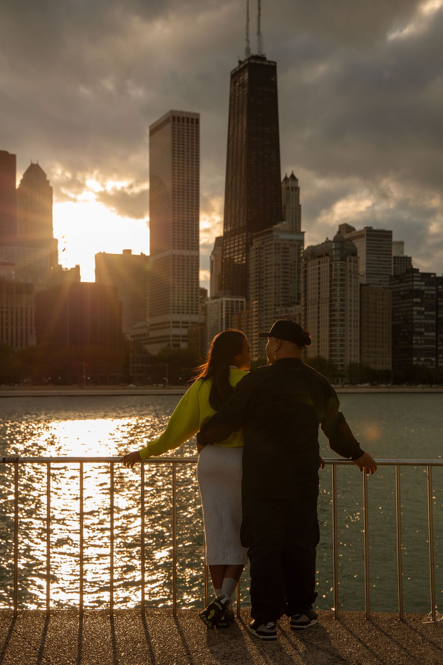 Couple embracing at sunset on Chicago waterfront with iconic skyline and John Hancock Center in background.