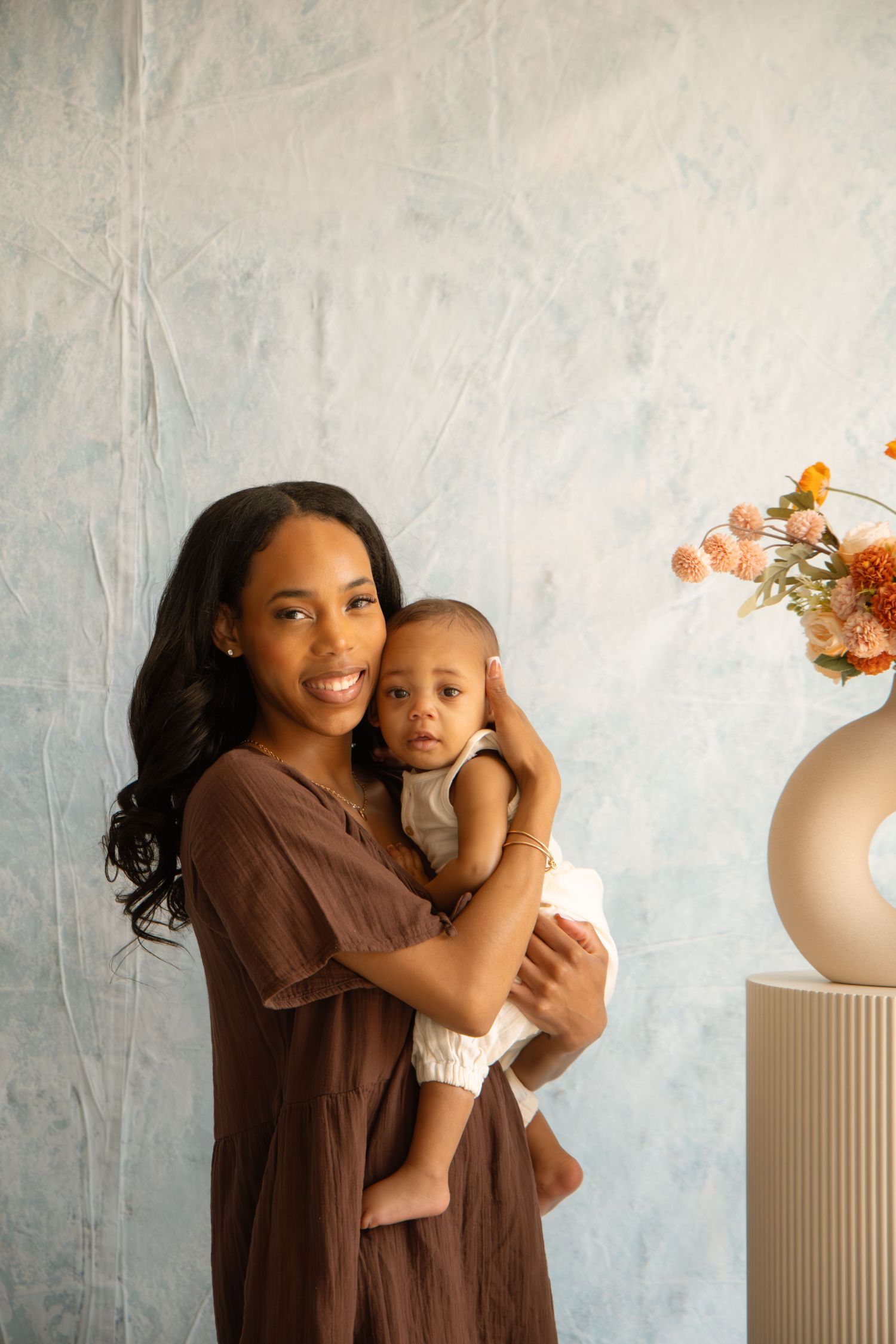 A smiling mother in a brown dress holds her baby near a floral arrangement against a blue textured wall.
