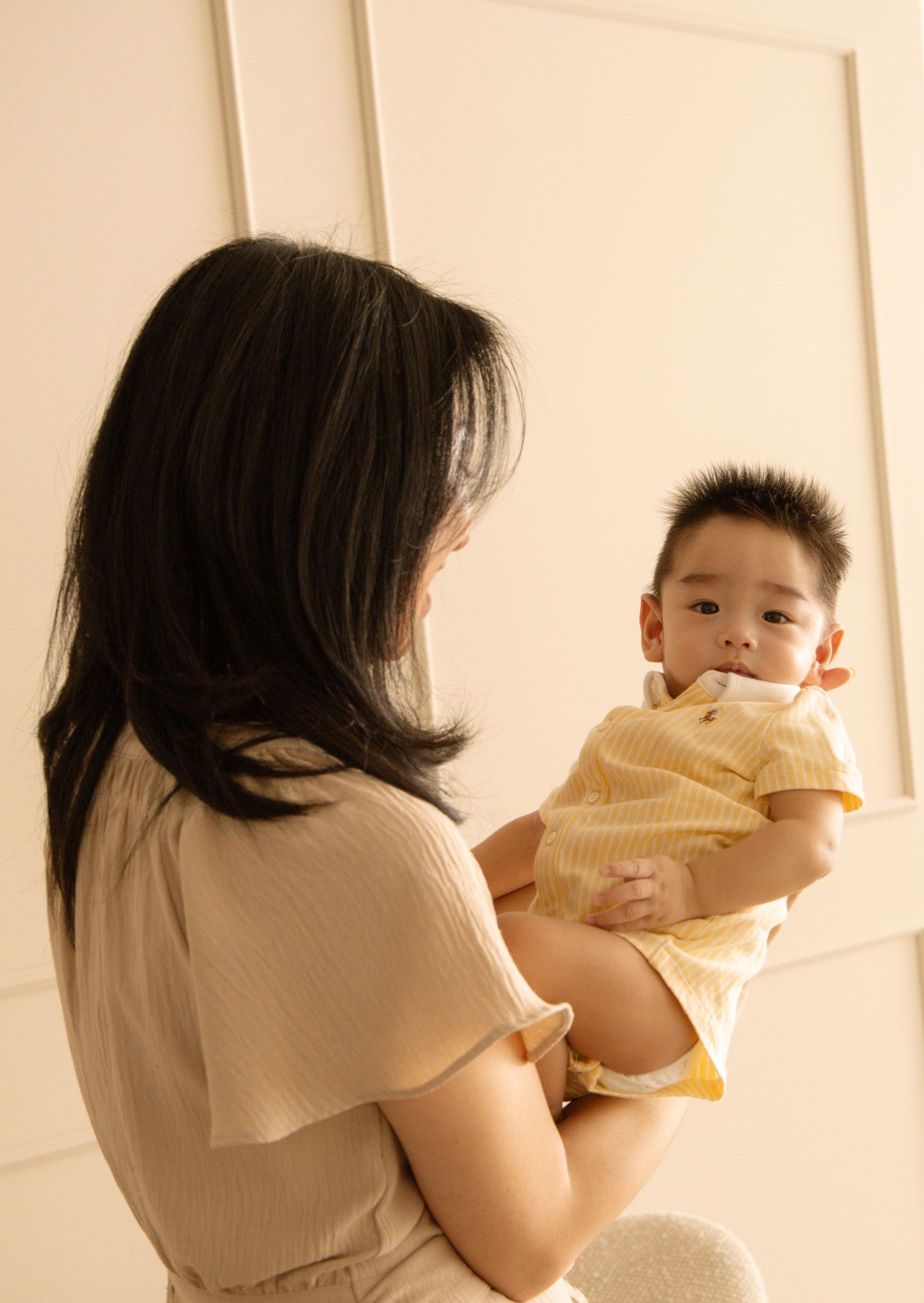 A mother holds her baby boy dressed in yellow, facing each other in a bright, softly lit room.