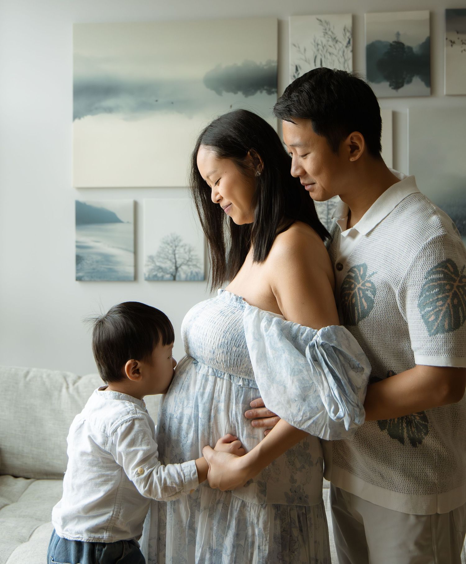A pregnant Asian woman in a blue dress poses with her husband and toddler son touching her belly in a bright room.