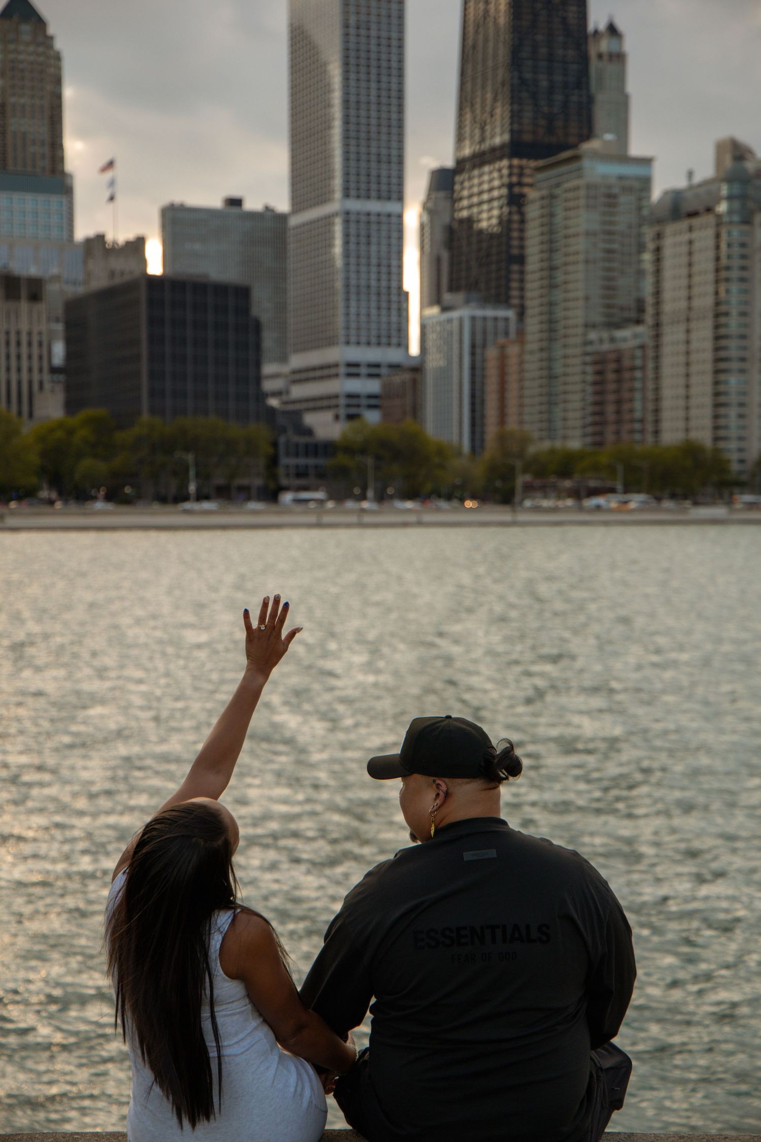 Couple sitting by waterfront admiring Chicago city skyline, woman raising hand, man in Essentials black jacket.