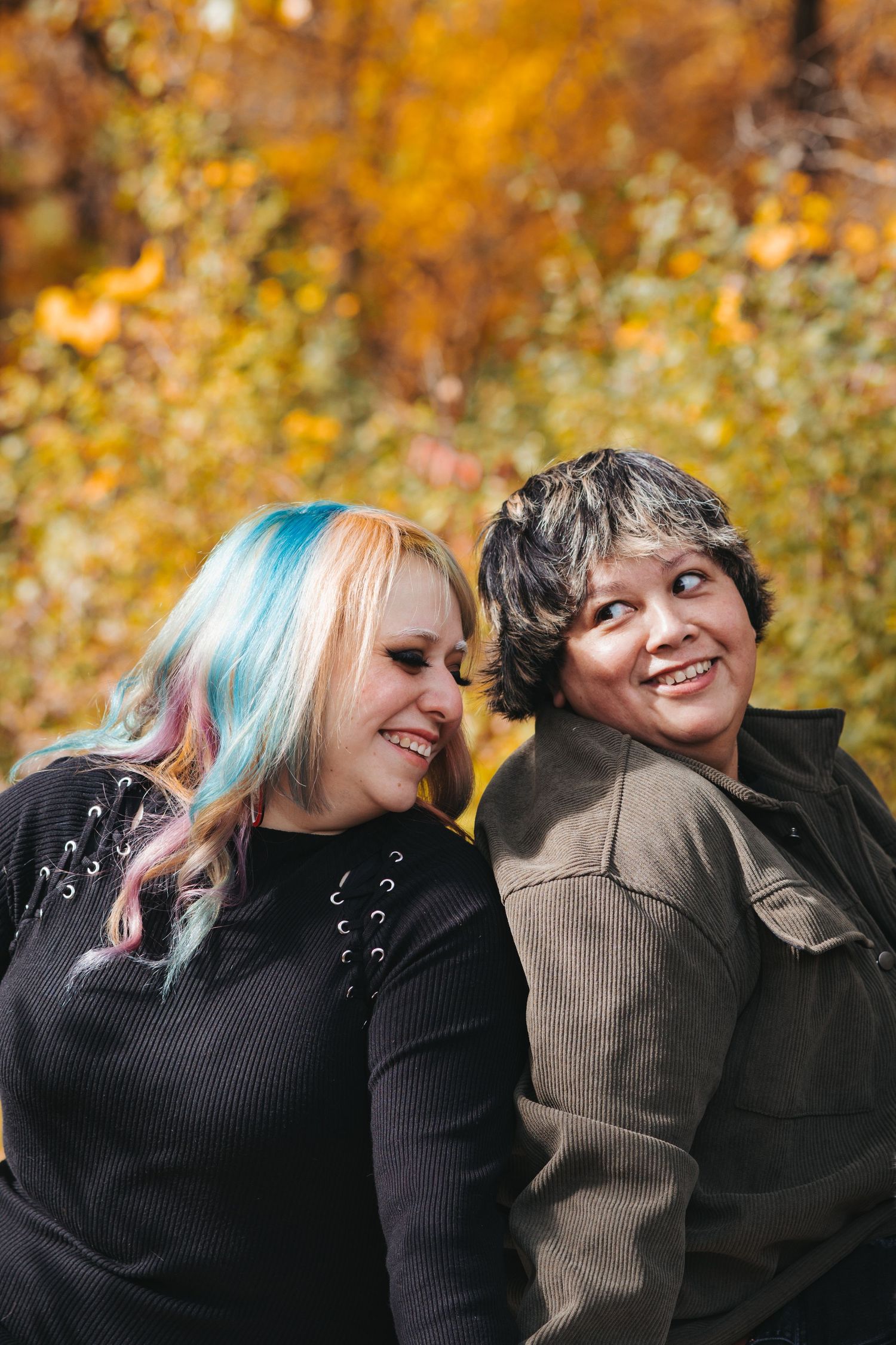 Two women with colorful and gray hair laughing together against vibrant autumn foliage background.